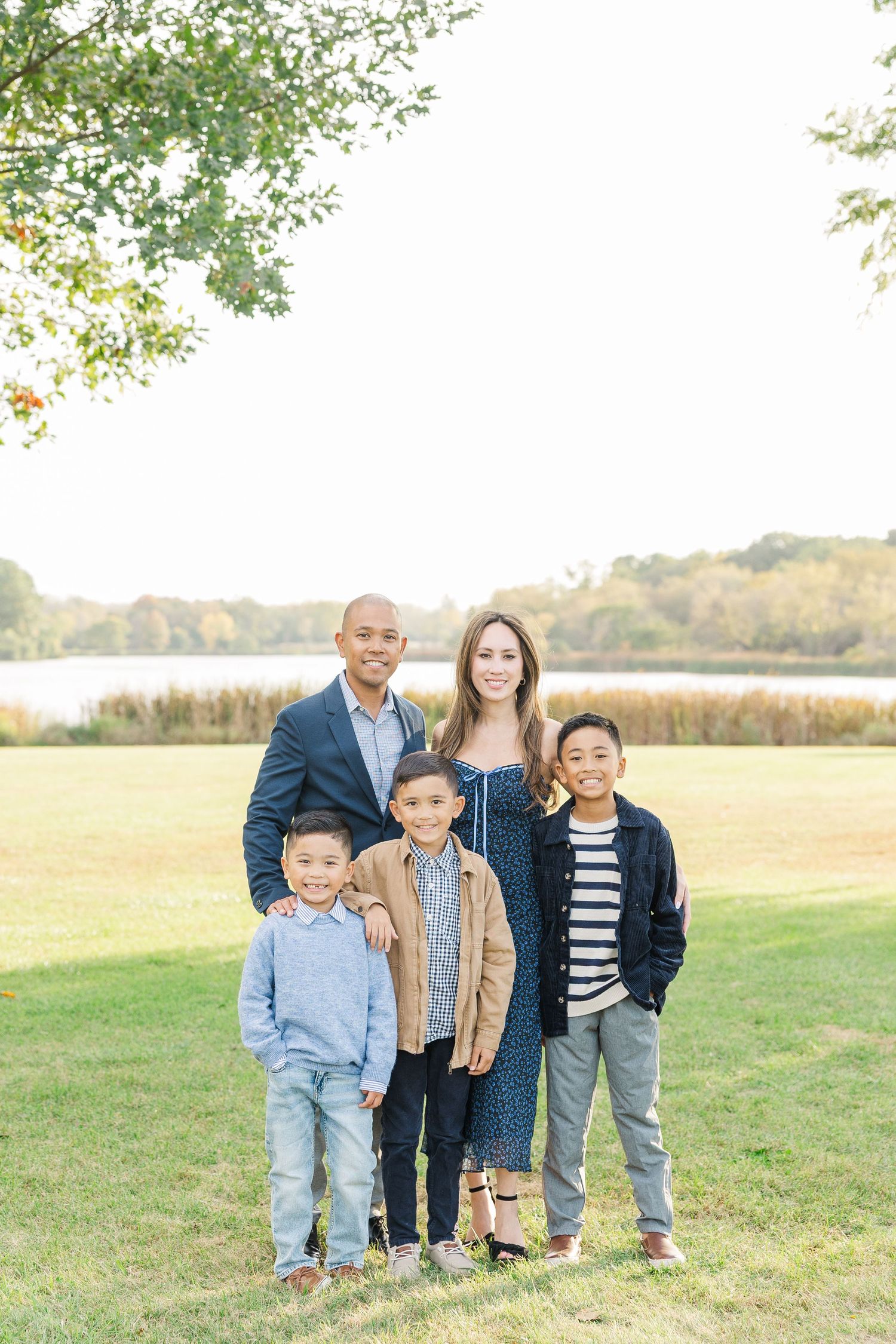 A family stands together outdoors on grass with a lake and autumn trees visible in the background. Busse Woods