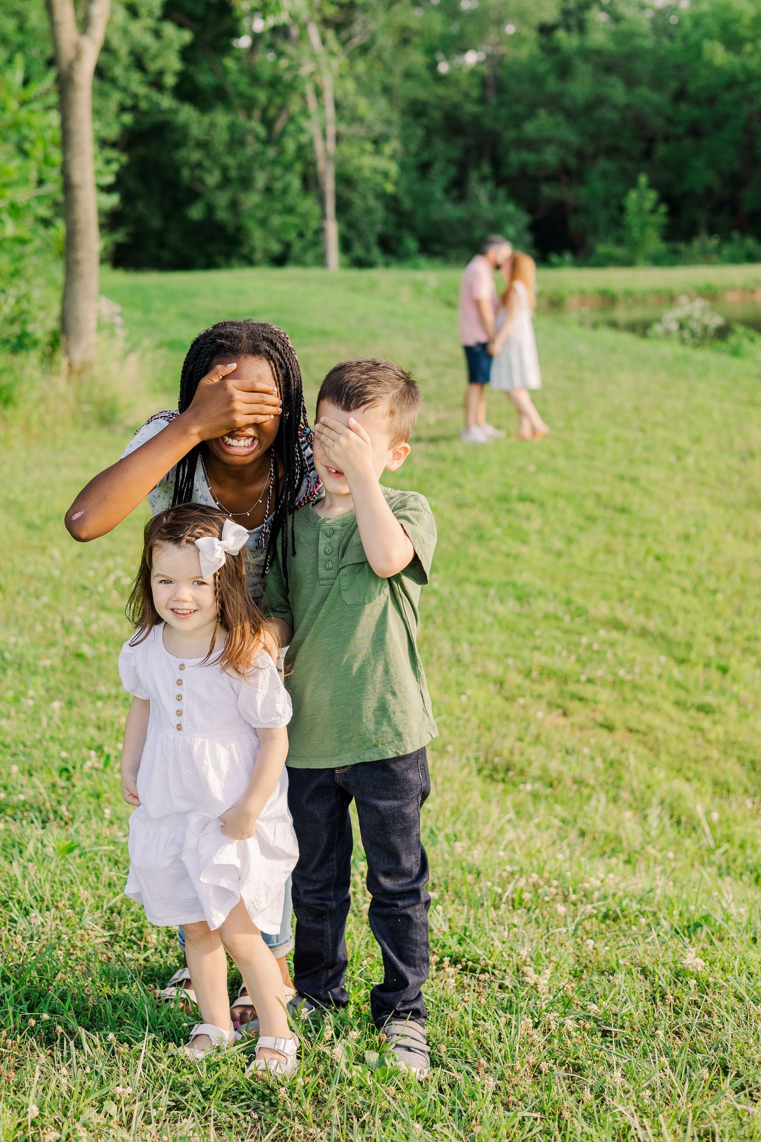 Children and caregiver playing hide and seek in a sunny park with people in the background.