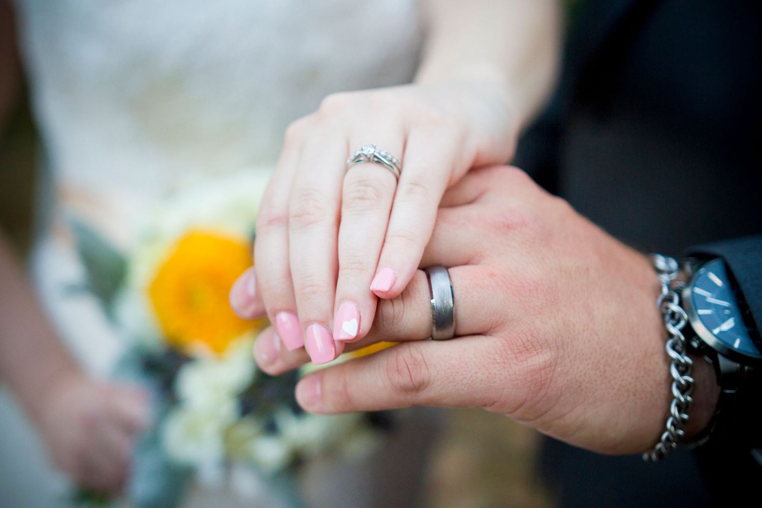 Close up of newlywed couple's hands showing wedding rings with flowers in background.