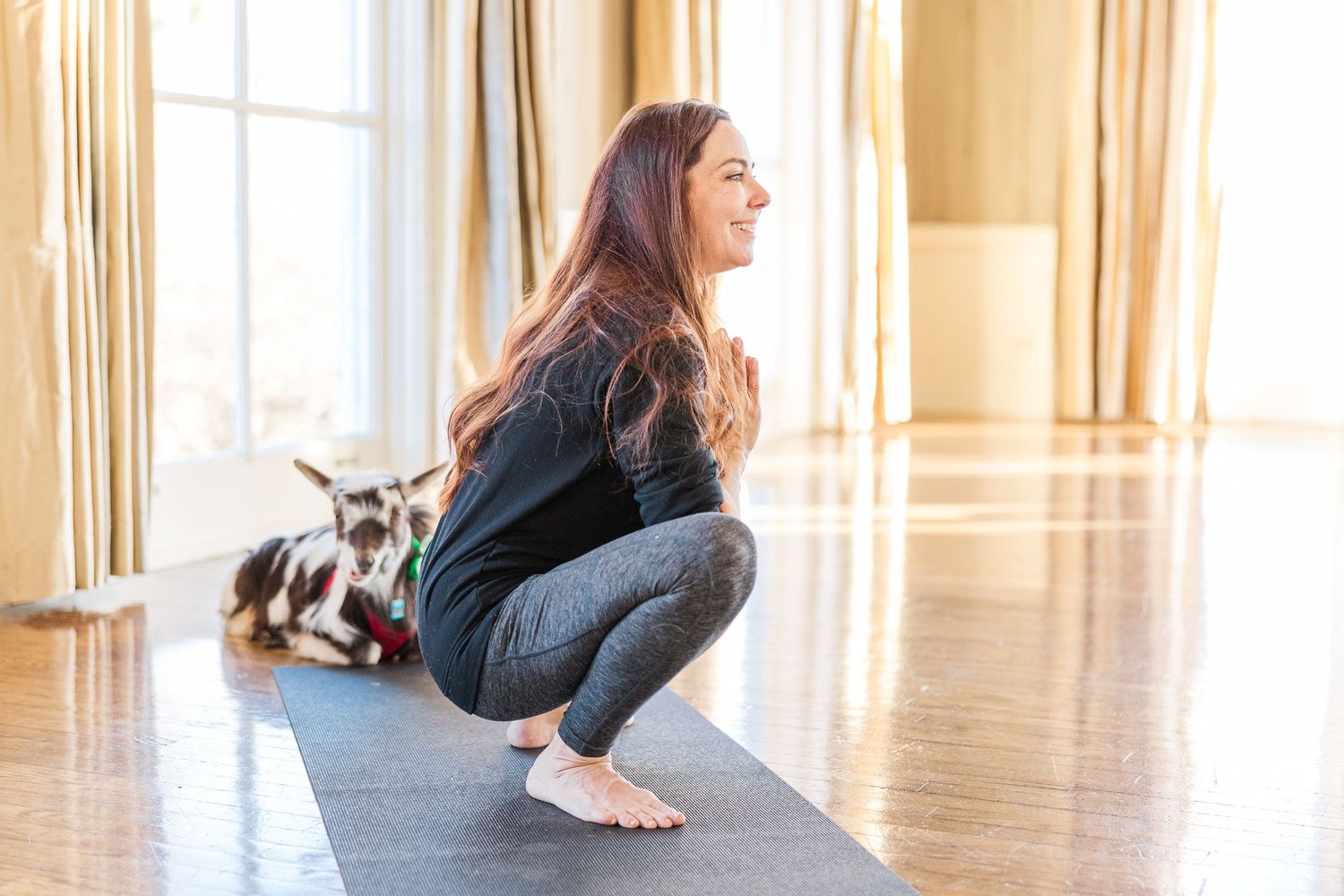 A person demonstrates proper squat form on a yoga mat while a spotted dog rests in the background.