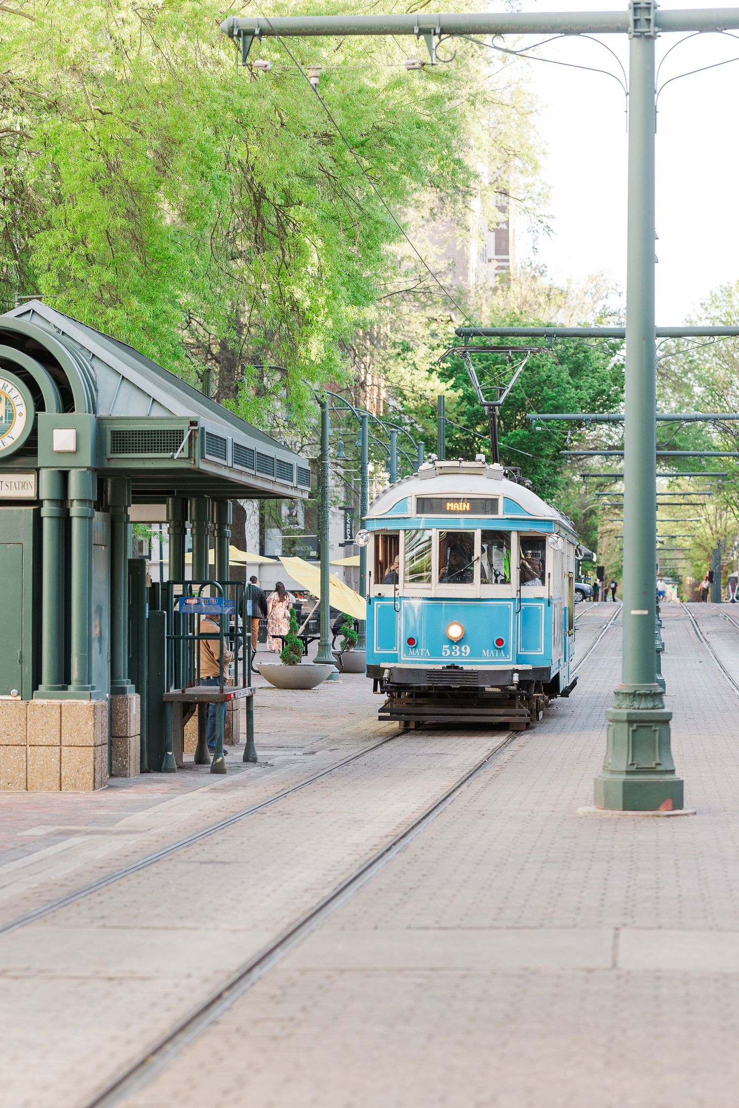 A vintage blue trolley car stops at a station along a tree-lined street.