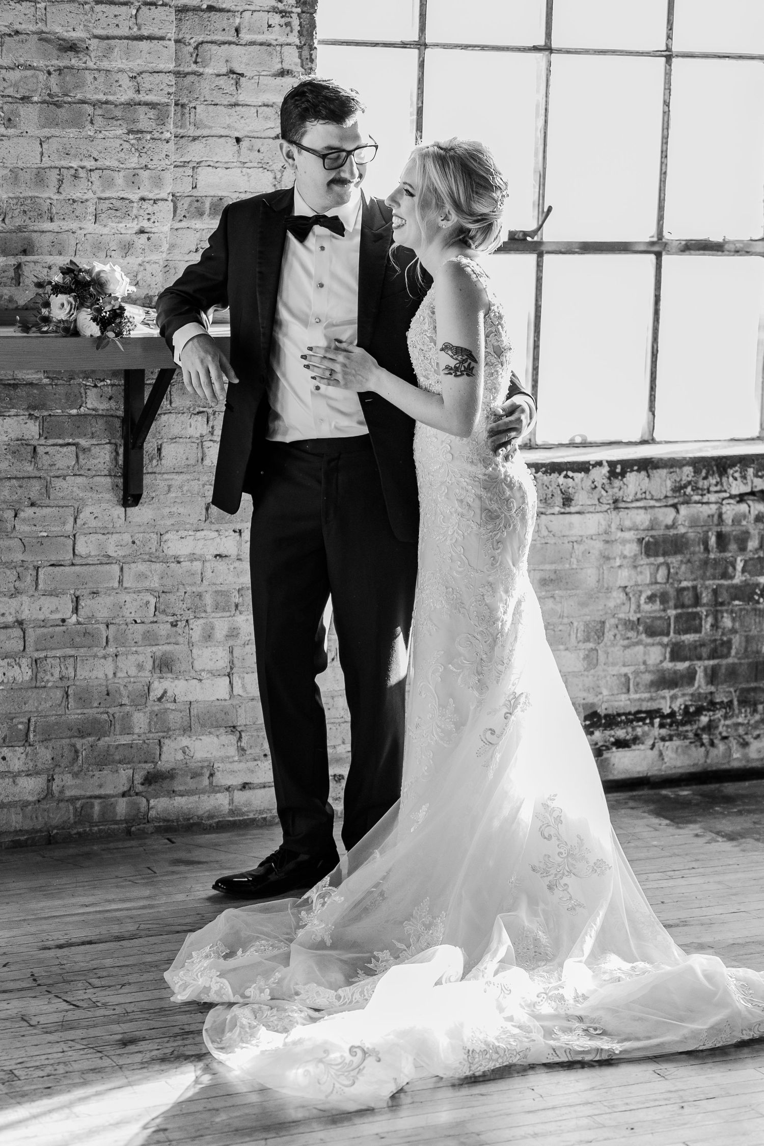 Black and white wedding photo with couple standing by large windows in a rustic brick venue.