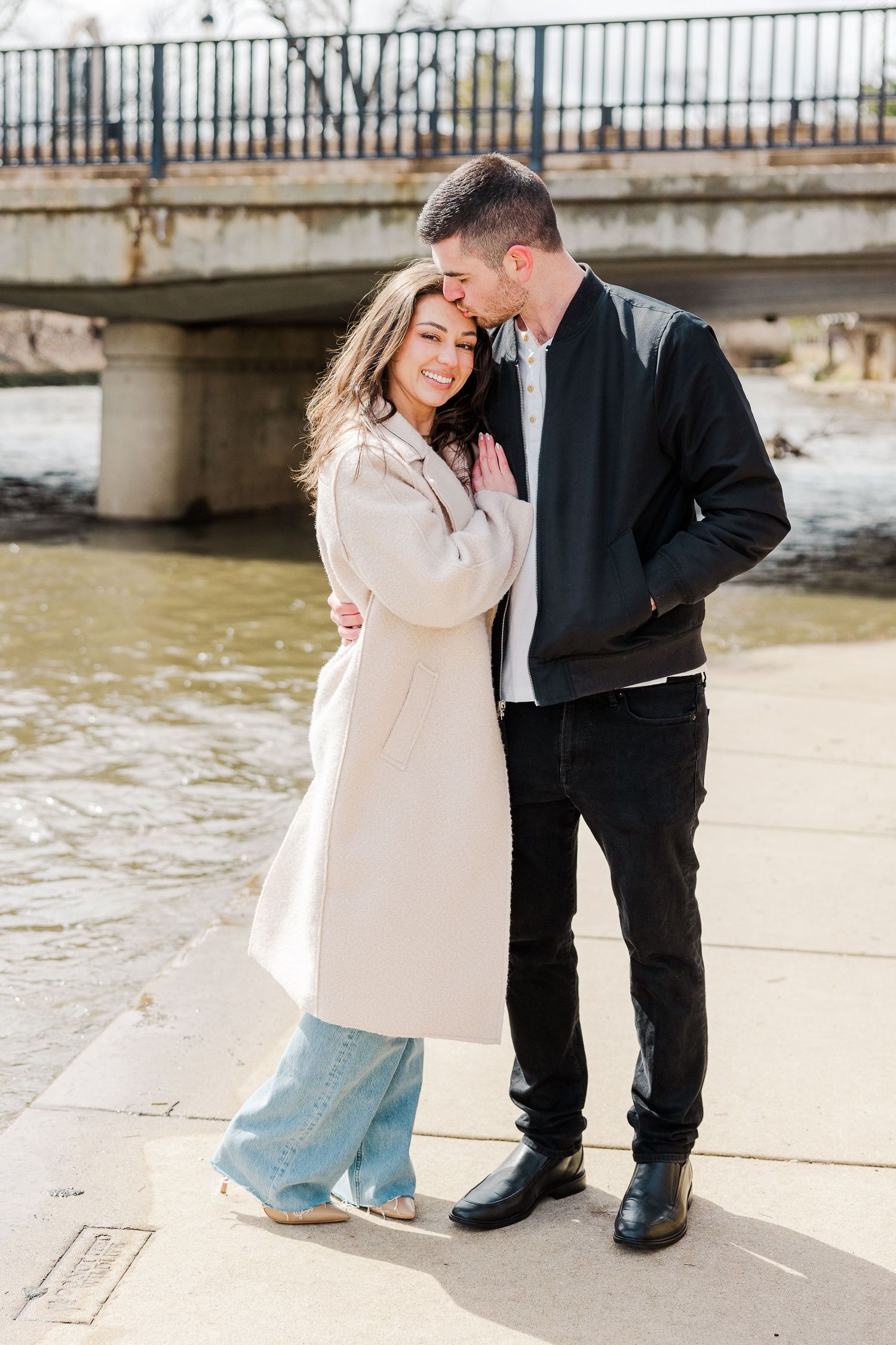 A couple embracing by a river with a bridge in the background on a sunny day.