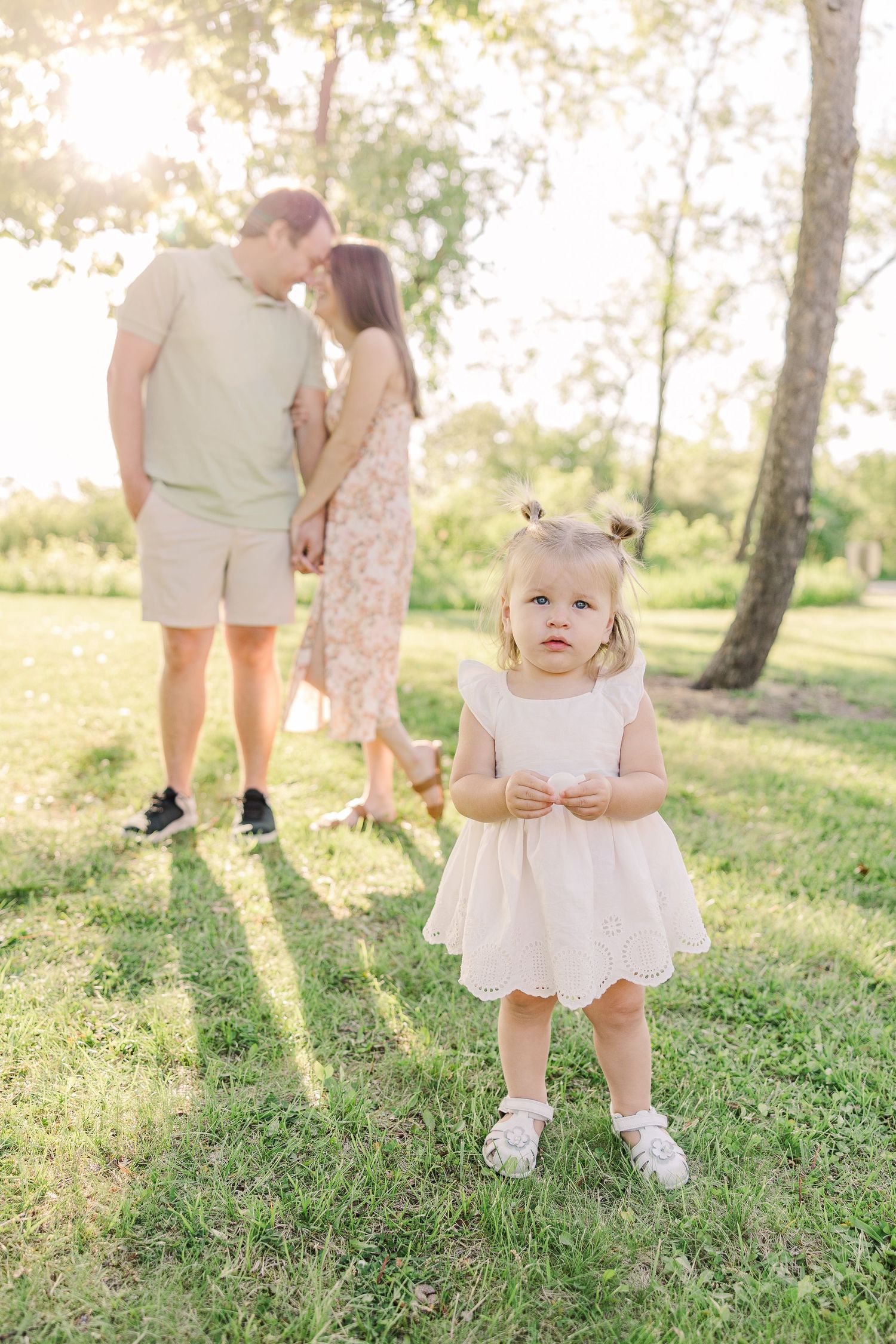 A sweet moment captured outdoors with a little one in a white dress under sunlit trees.