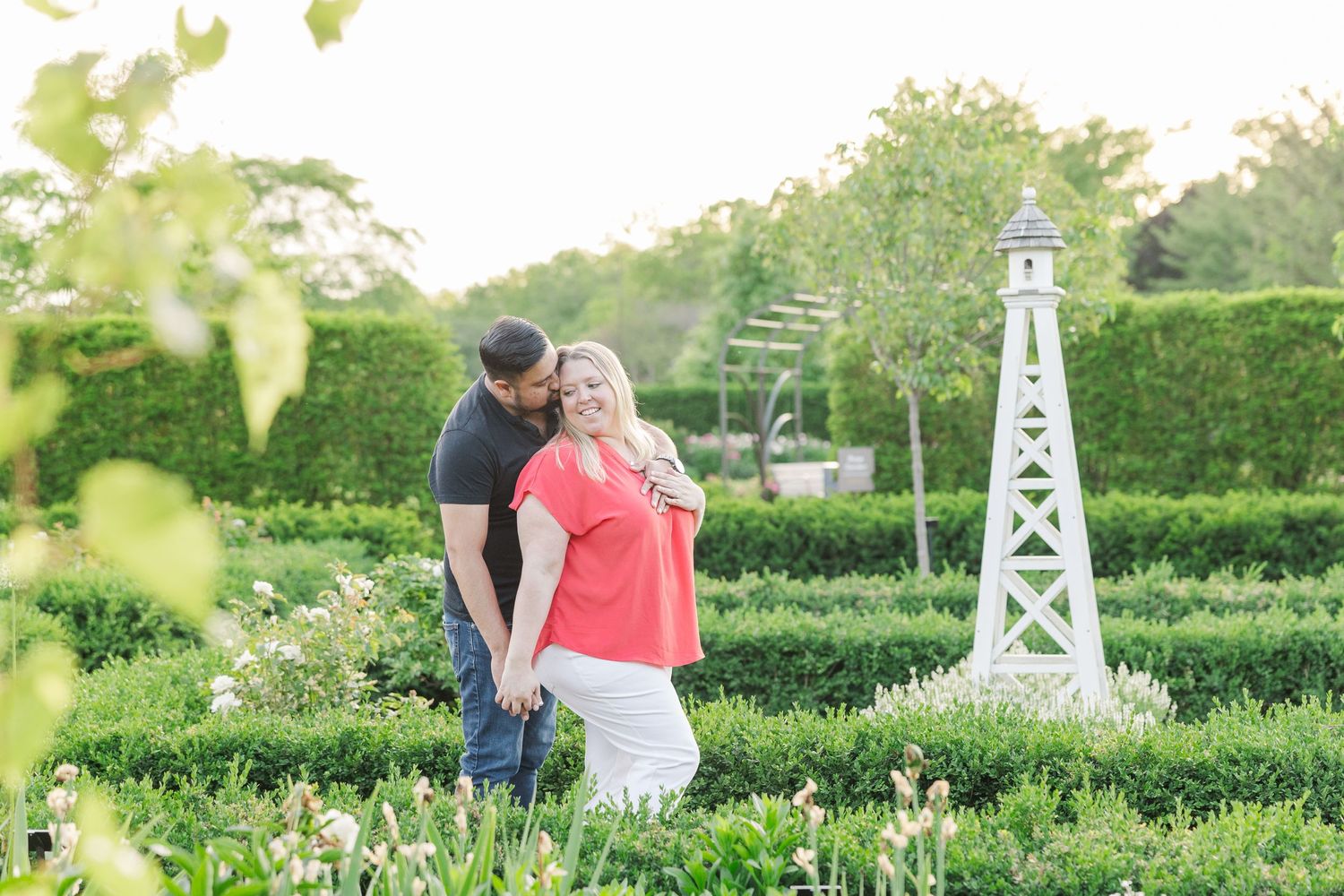 Couple embracing in a formal garden with white obelisk structure and well-maintained hedges.