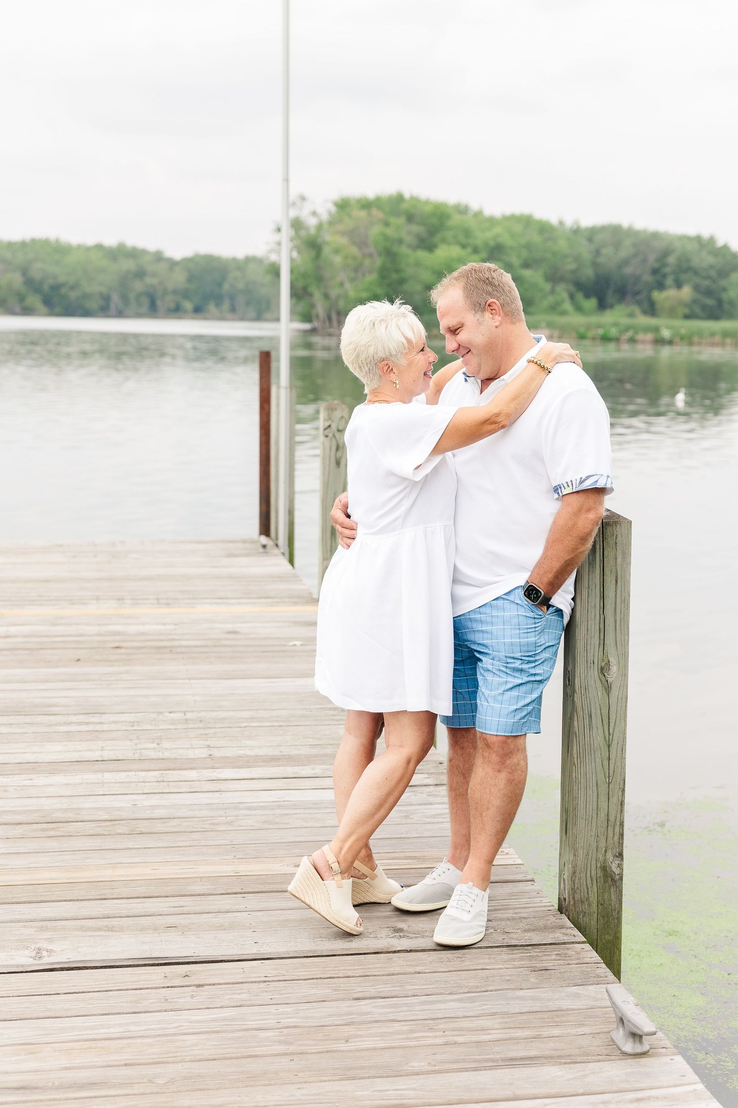 Couple in casual white outfits sharing romantic moment on wooden pier by lake.