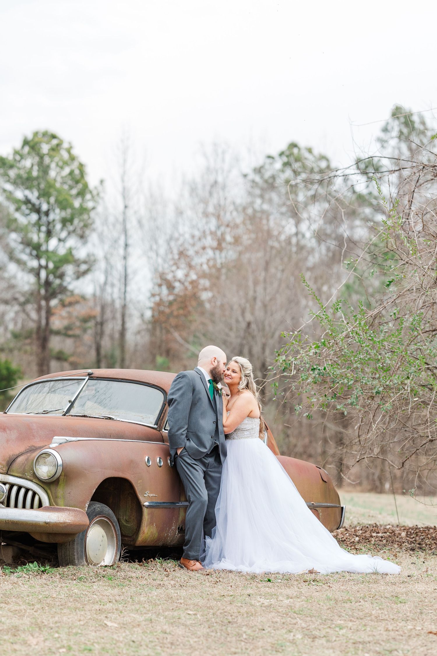 Romantic wedding photo with couple leaning against classic rusty automobile.