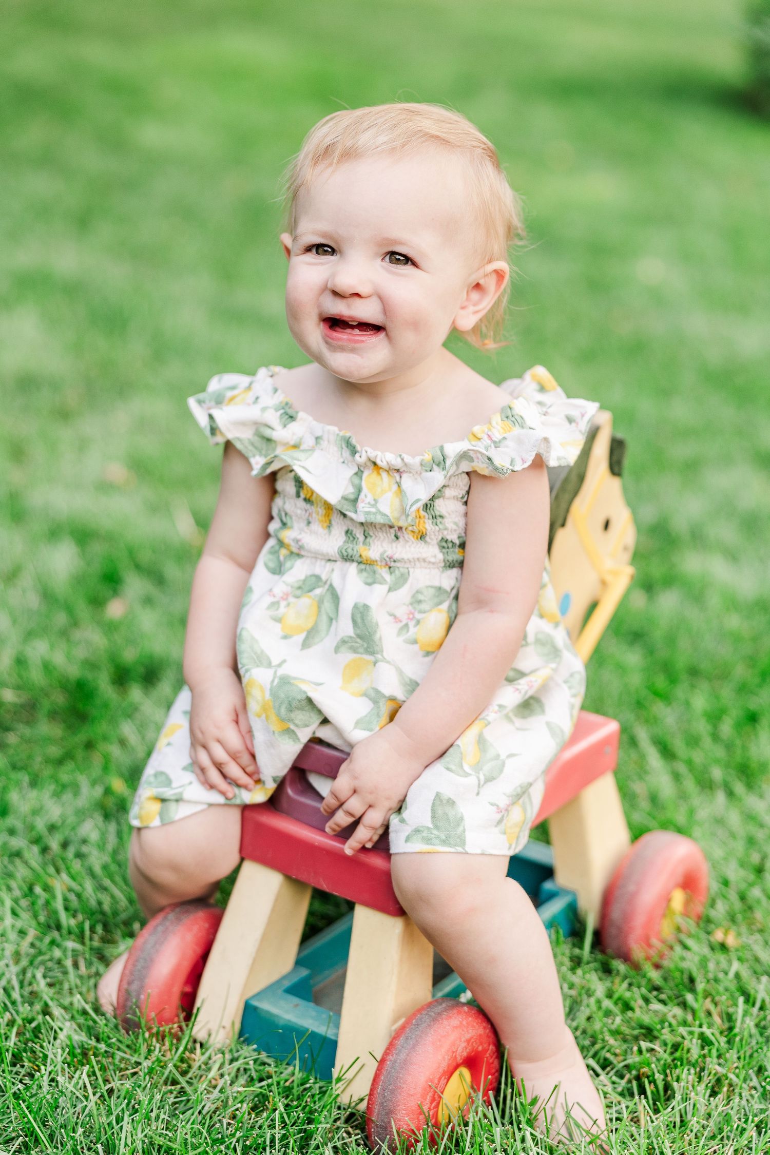 A joyful young child wearing a yellow patterned dress plays on a wooden riding toy on green grass.
