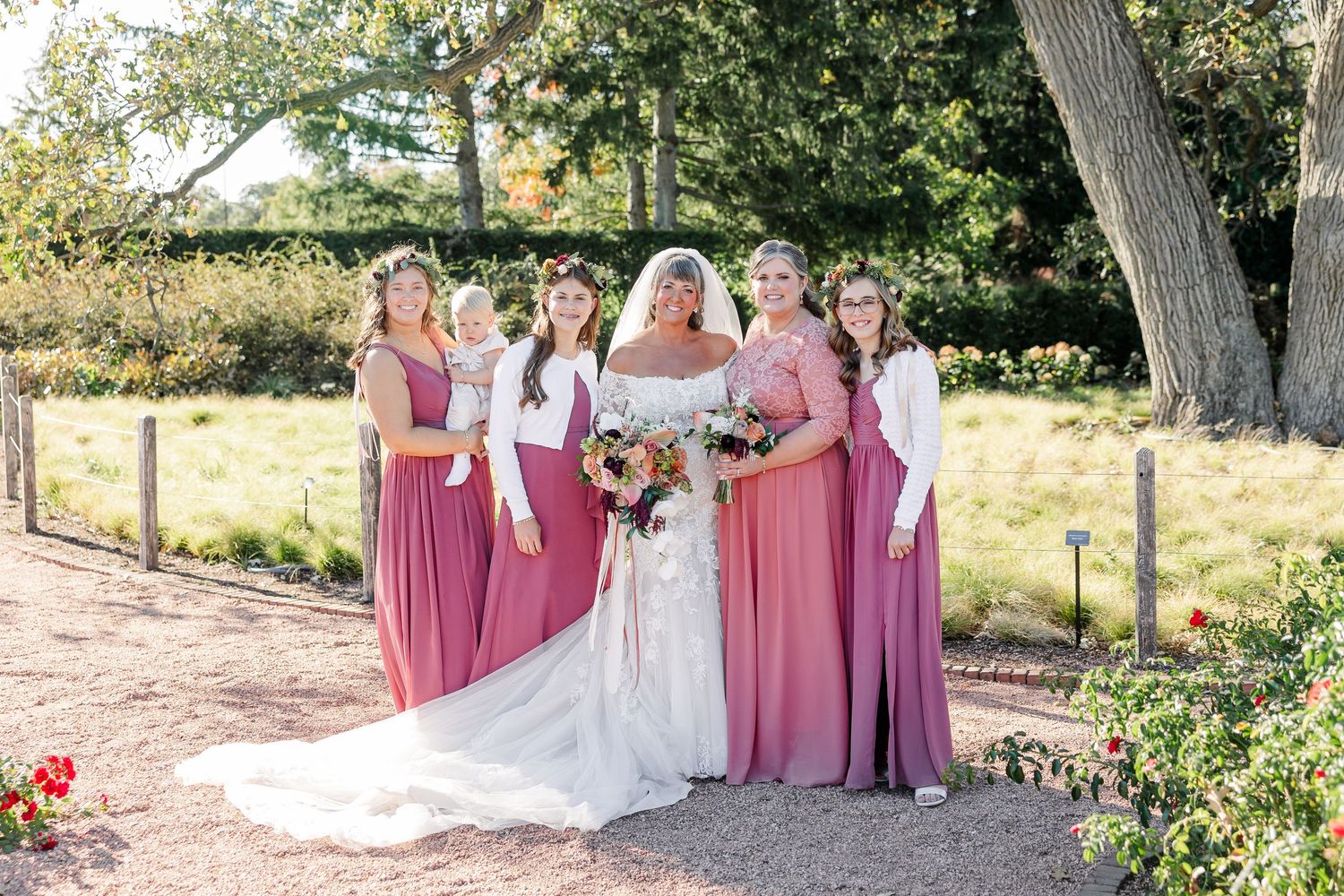 A bride in a white gown holding a bouquet poses with four bridesmaids in mauve dresses and a baby on a garden path beside roses.
