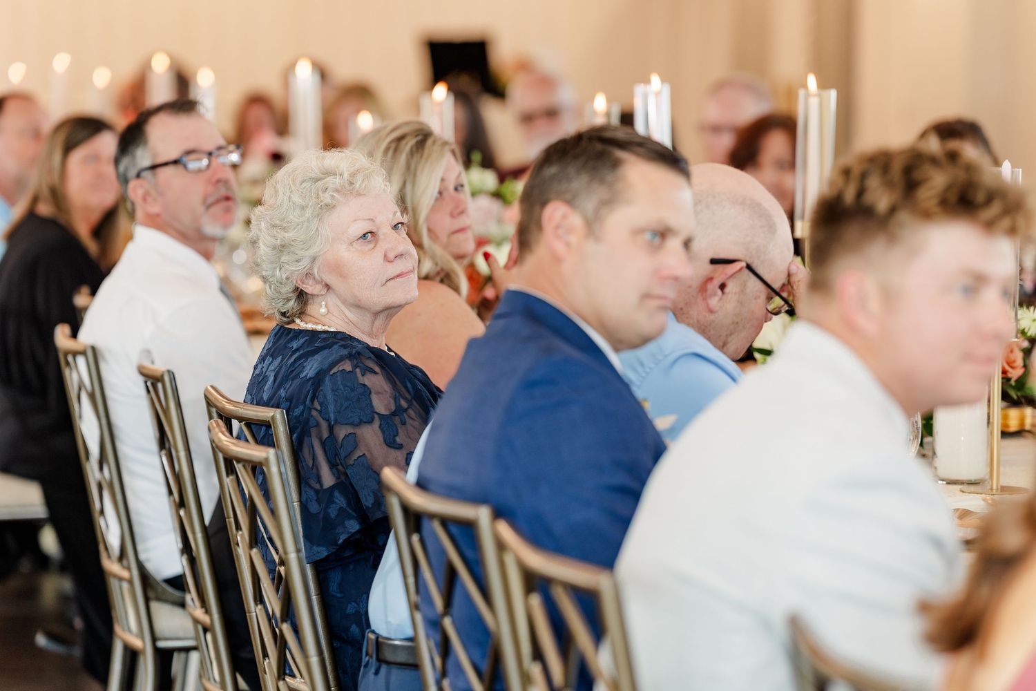 Wedding guests seated at a reception table listen attentively, with glowing candles and floral centerpieces in the background.