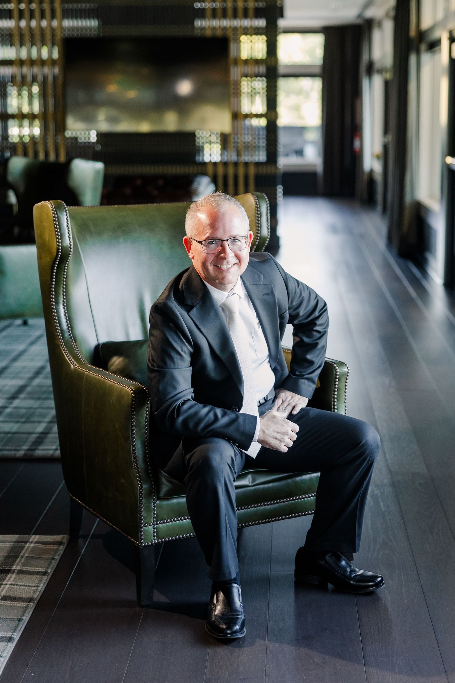 Smiling man in glasses, dark suit and tie sits in a green leather armchair in an elegant lounge with dark wood floors.
