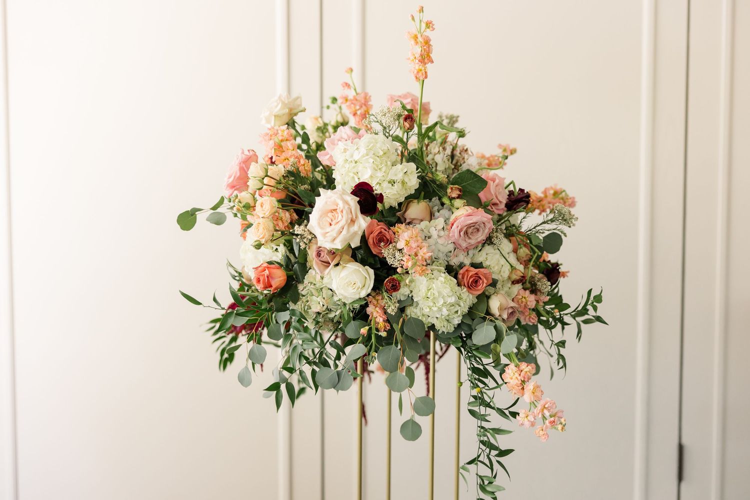 Tall wedding floral centerpiece with peach and blush roses, white hydrangea, and trailing greenery on a stand.