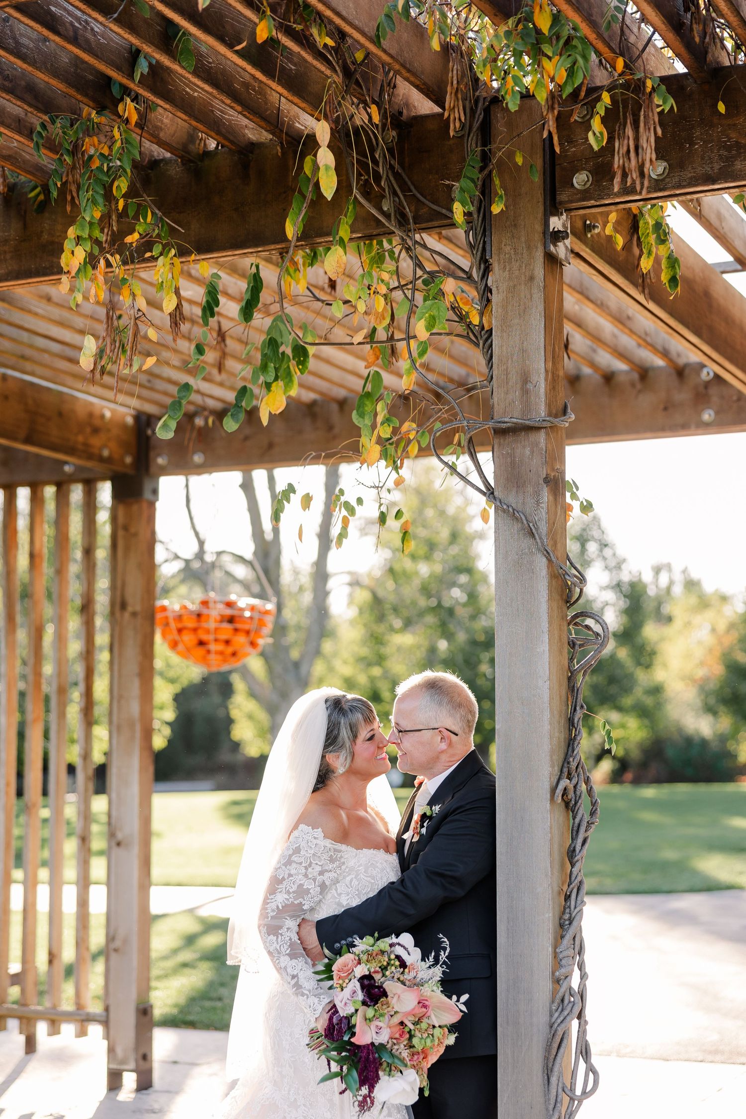 Bride and groom embrace under a rustic wooden pergola draped with autumn vines, she holding a pink and burgundy floral bouquet.