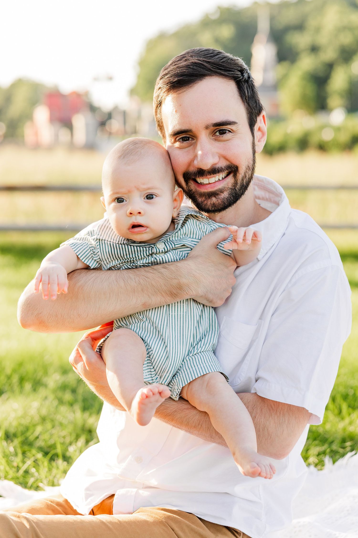 Smiling father holding his baby boy while sitting on grass in a sunny outdoor setting.
