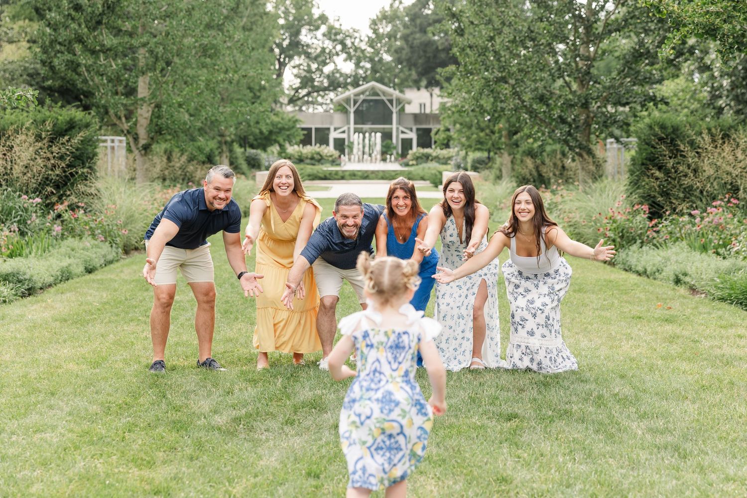 Family of seven smiling and reaching toward a toddler on a green lawn in front of an elegant garden estate.