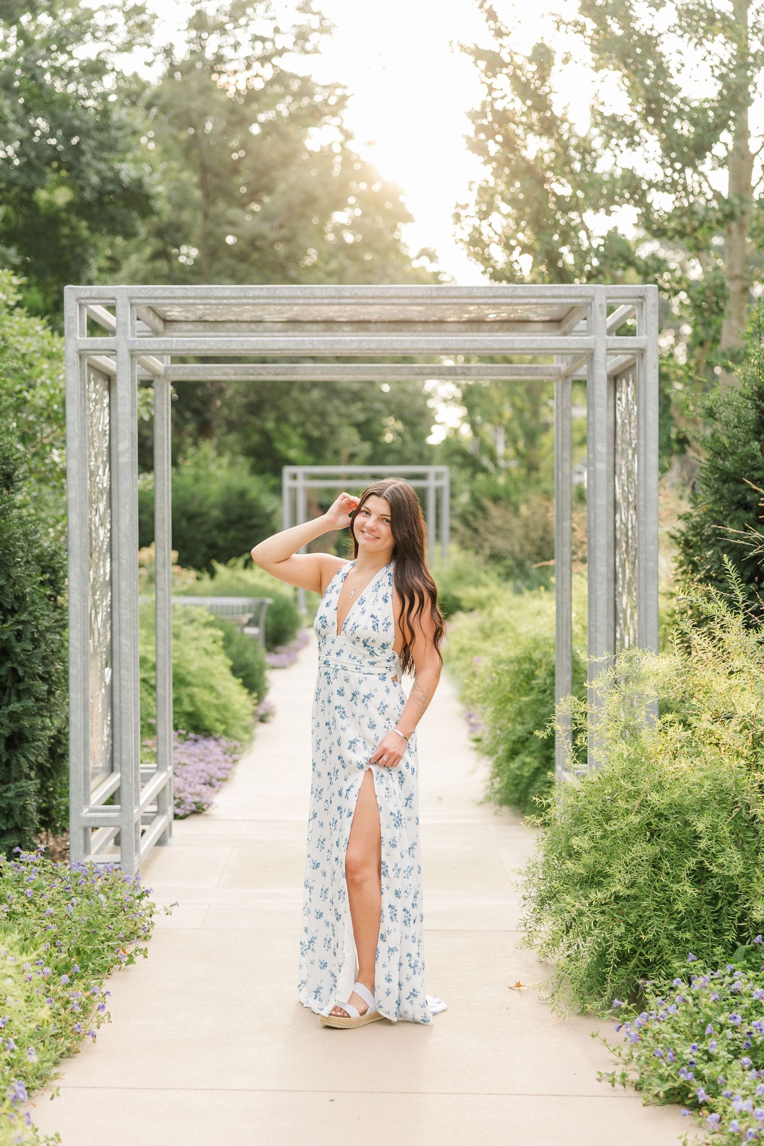 Young woman in floral maxi dress posing in a garden pergola pathway surrounded by lush greenery and soft sunlight.