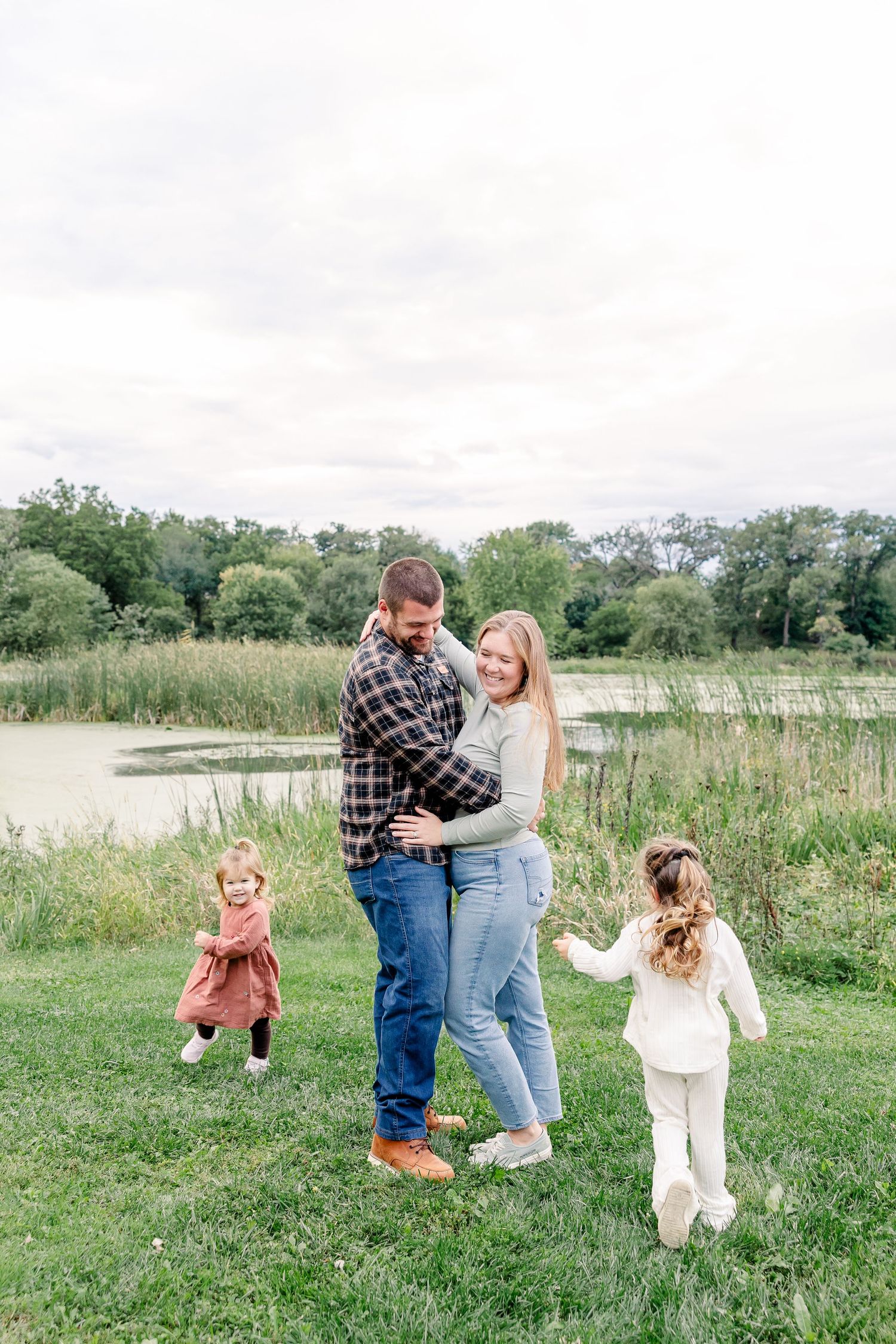 A happy family of four embraces and laughs together near a scenic pond surrounded by tall grass.