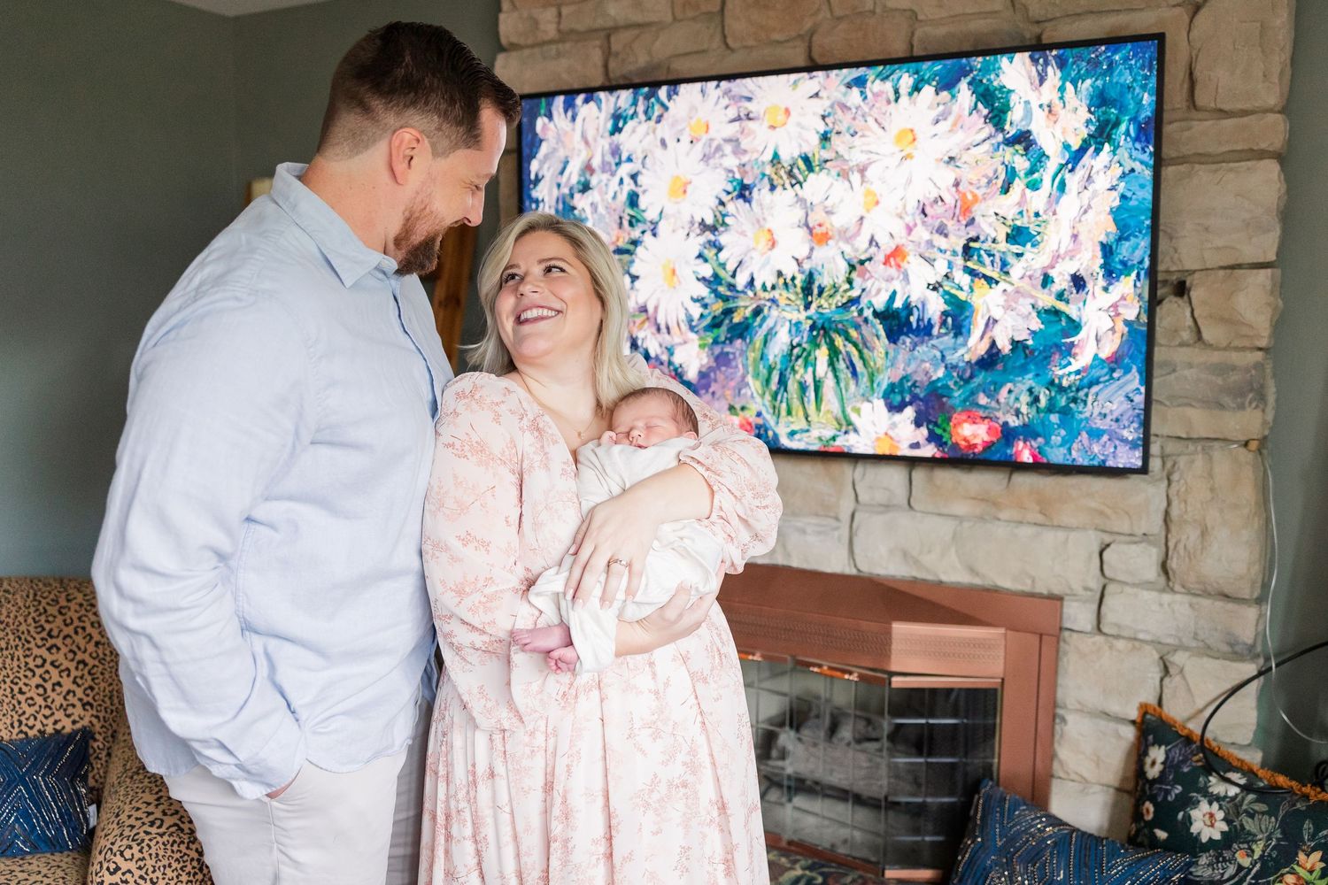 New parents in coordinating blue and pink outfits admiring their newborn baby in front of stone fireplace.
