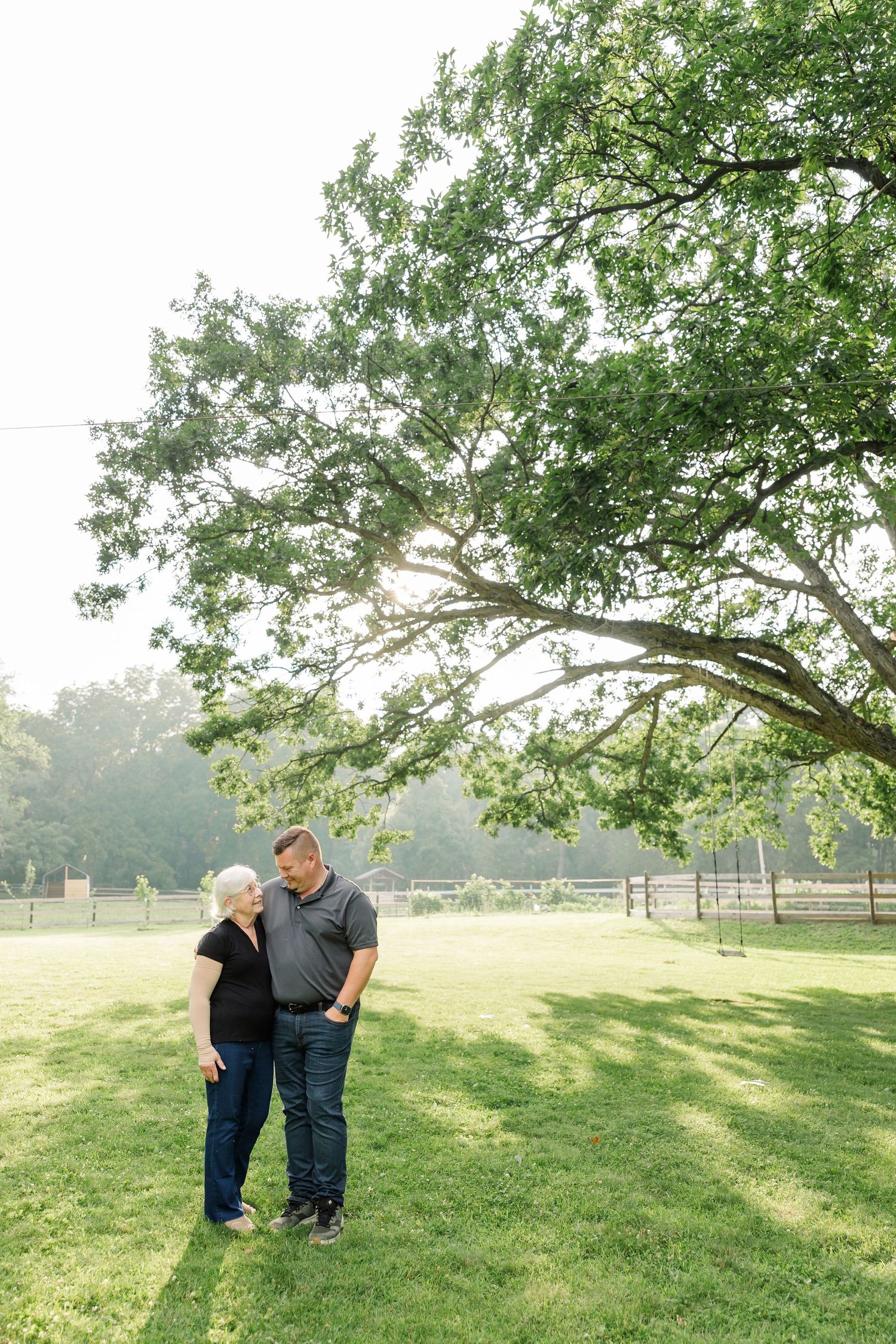 Couple standing close together on lush green grass beneath a large tree with sunlight filtering through.