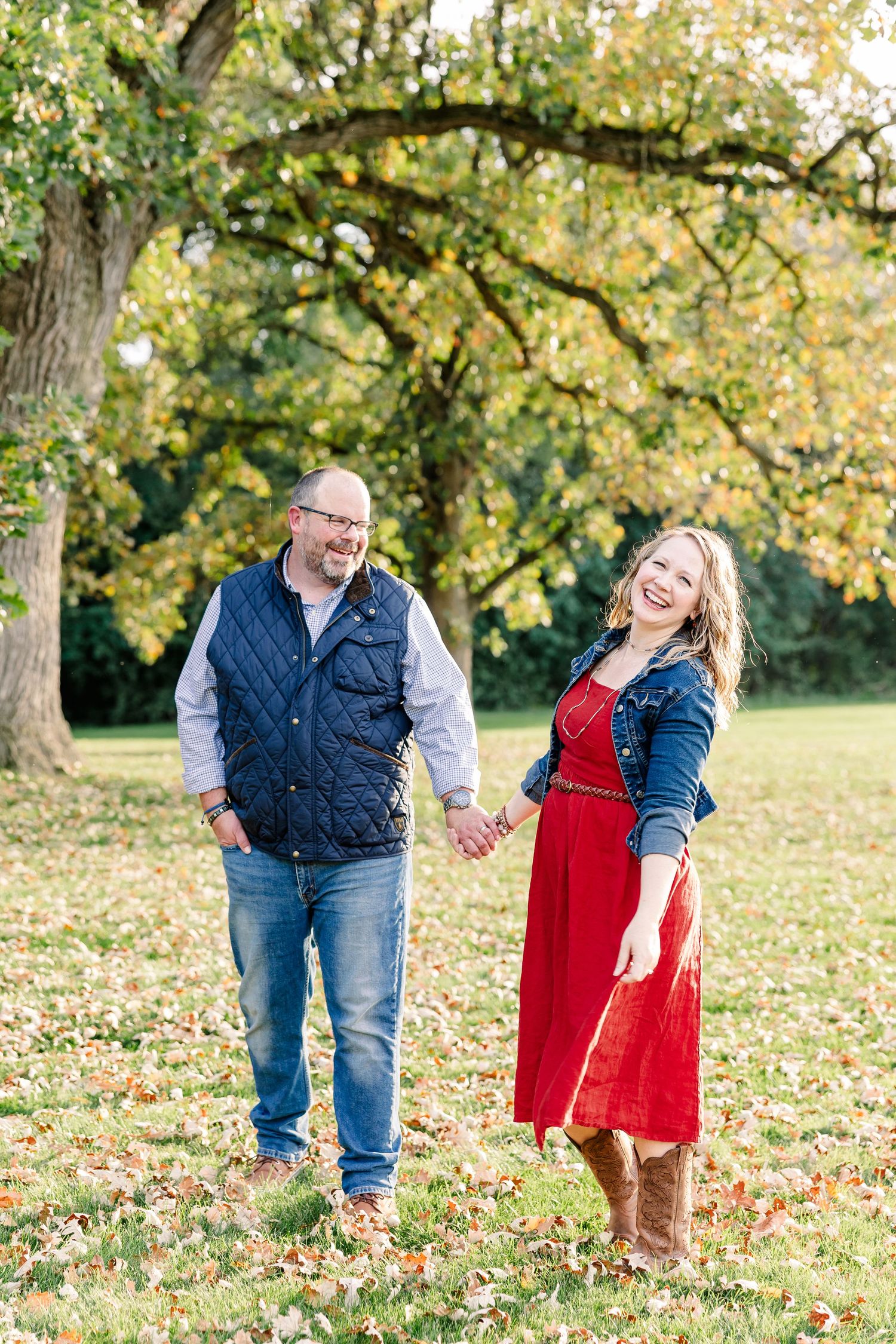 A laughing couple holds hands and walks across a leaf-covered lawn under an autumn tree, the woman wearing a red dress and boots.