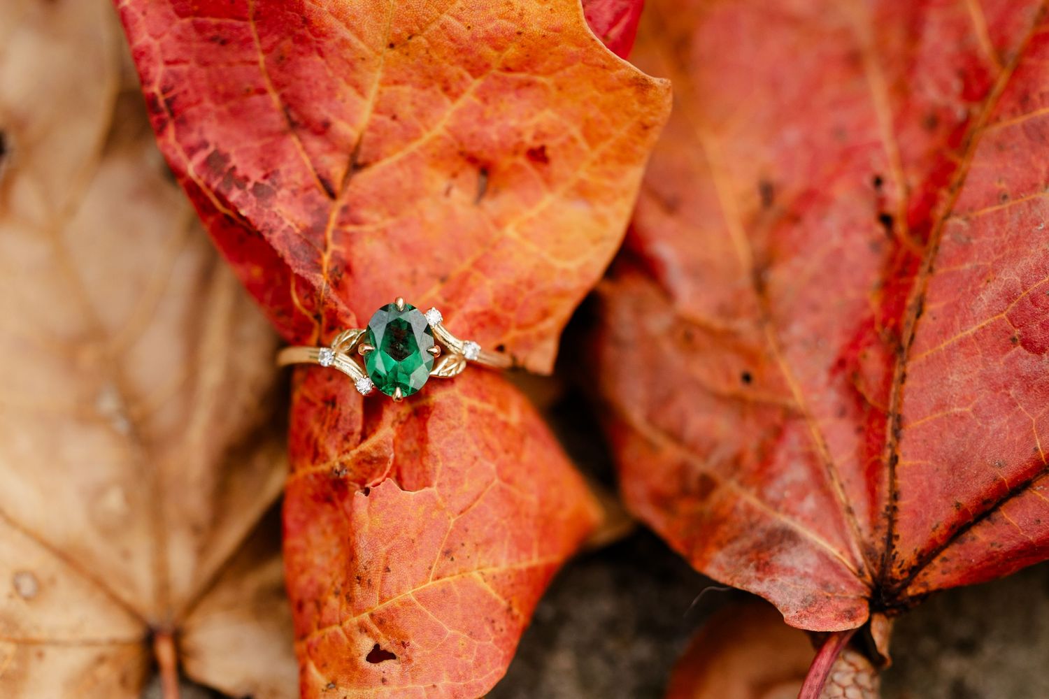 Emerald green gemstone ring with diamond accents resting on vibrant red and orange autumn maple leaves.