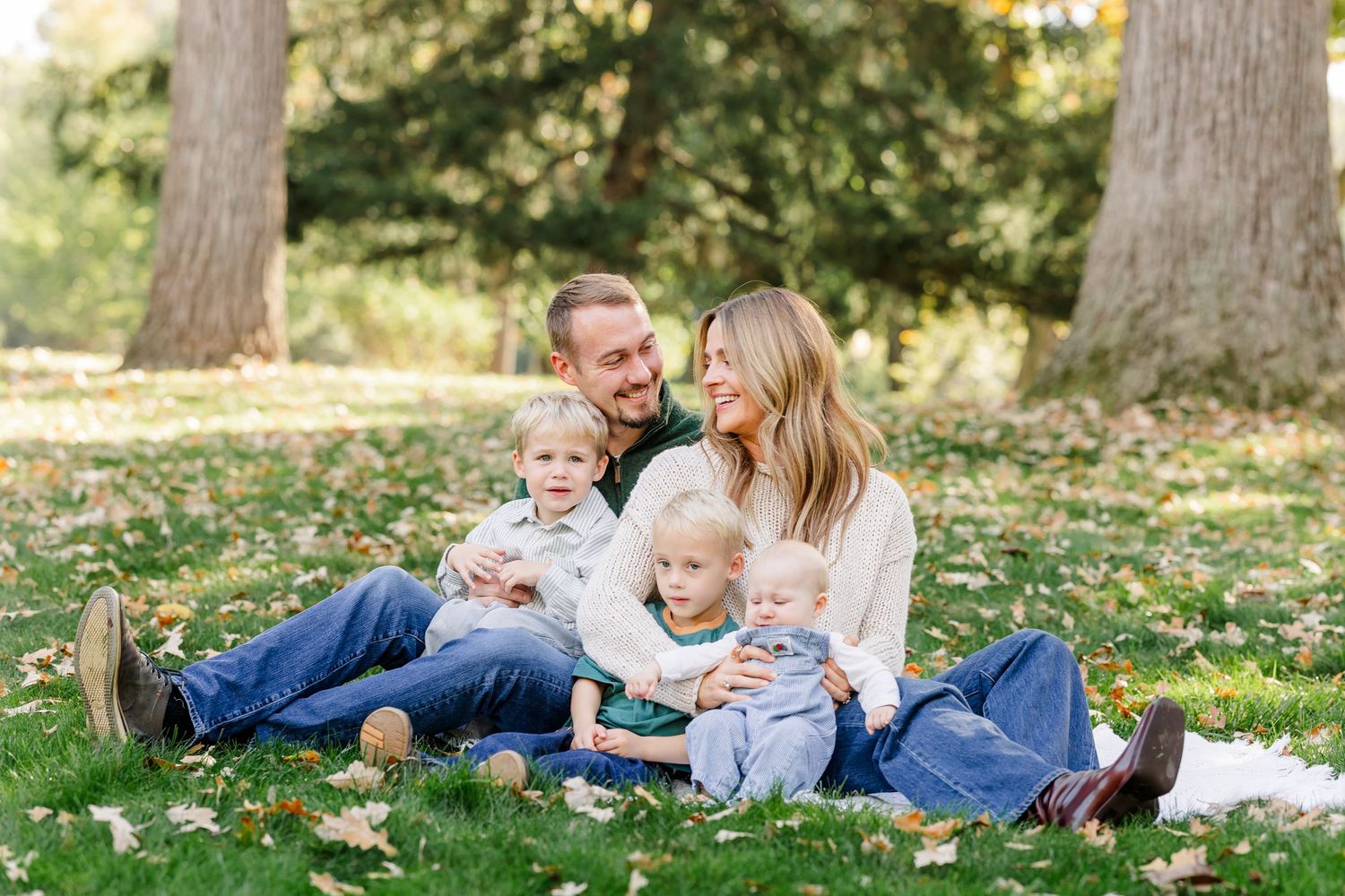 Happy family of four sitting on grass in a park during autumn, parents smiling with two young children.