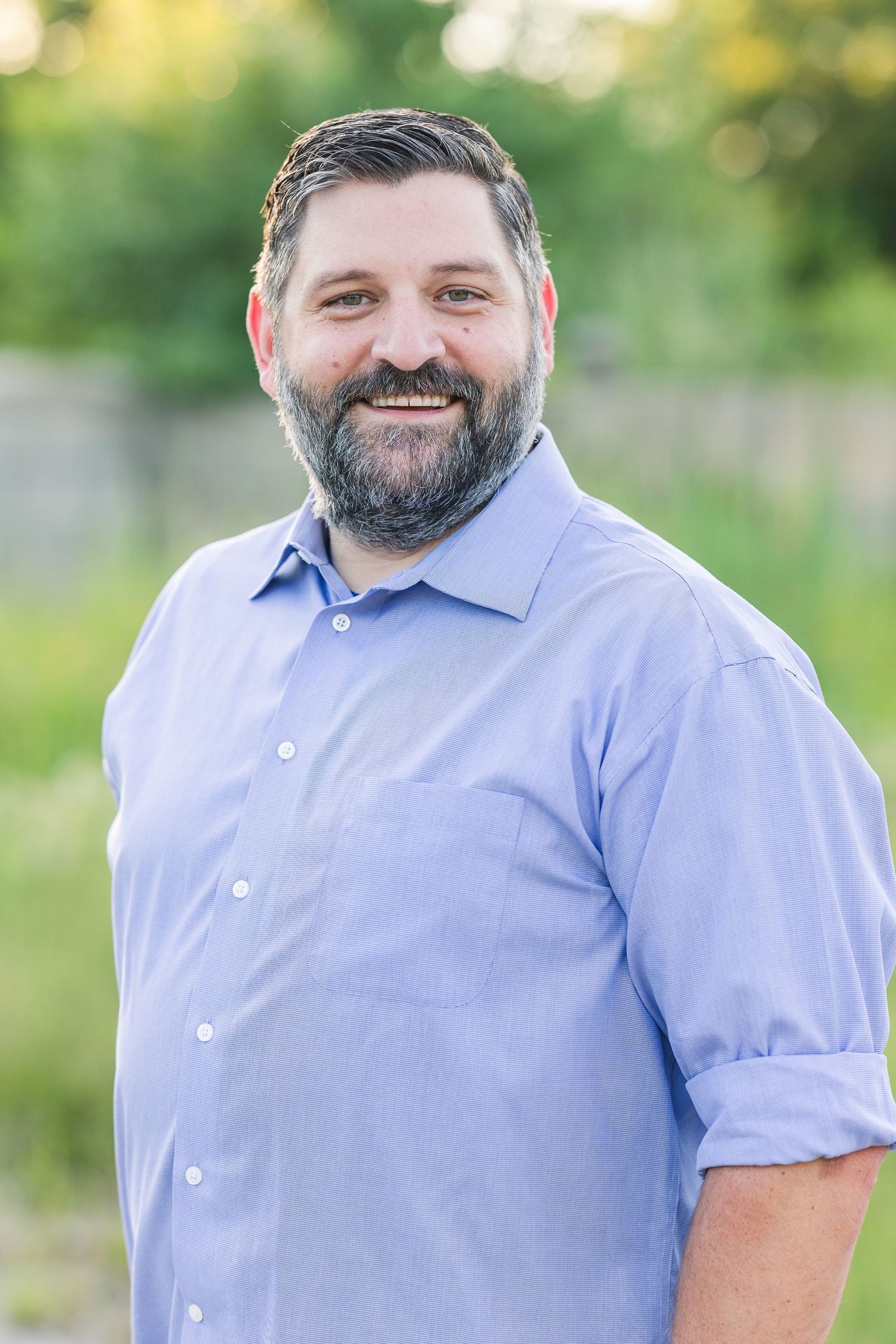 A person in a light blue button-down shirt smiles warmly while posing outdoors against a blurred natural background.