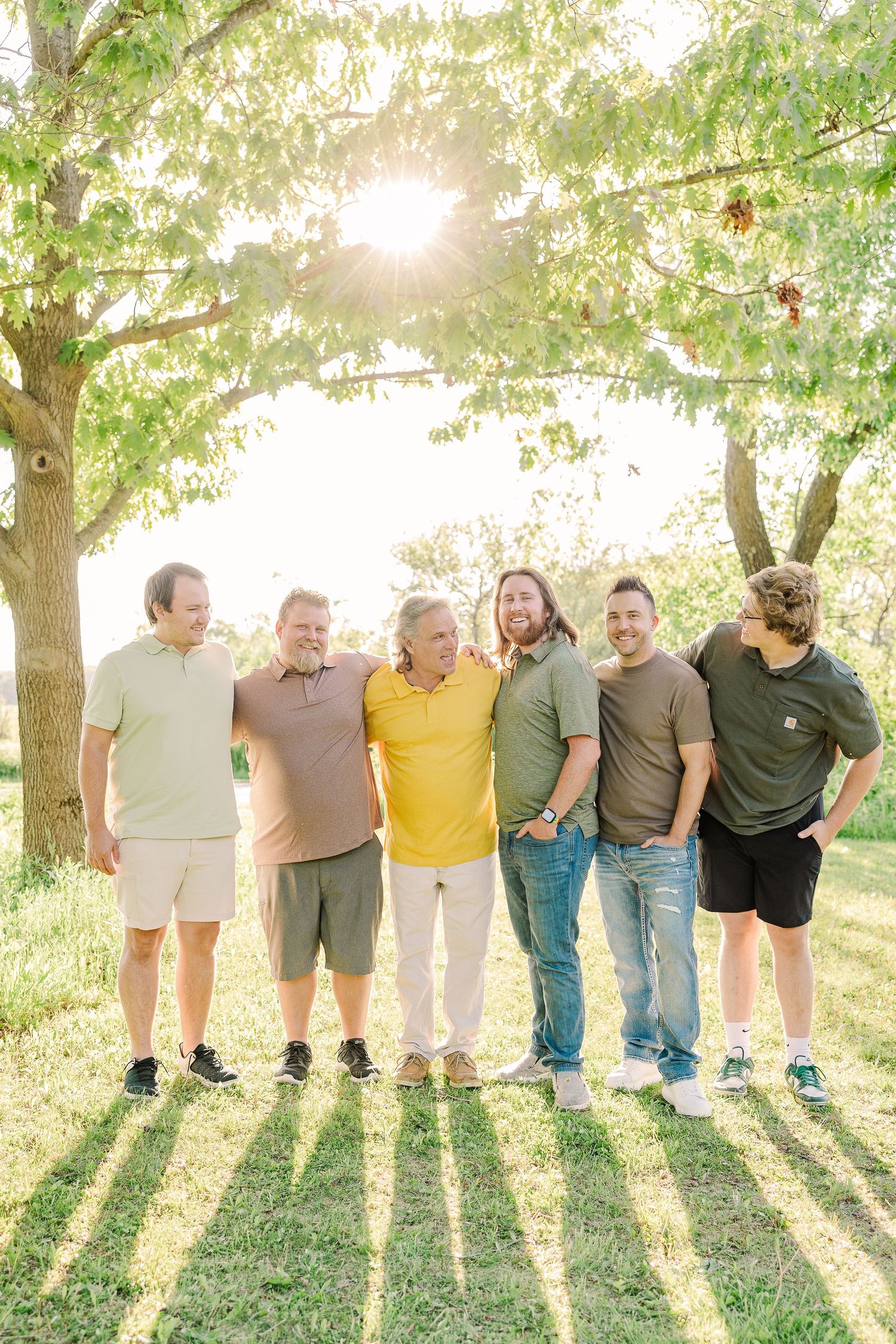 Group portrait of six people standing together and smiling under a sunlit tree casting long shadows.