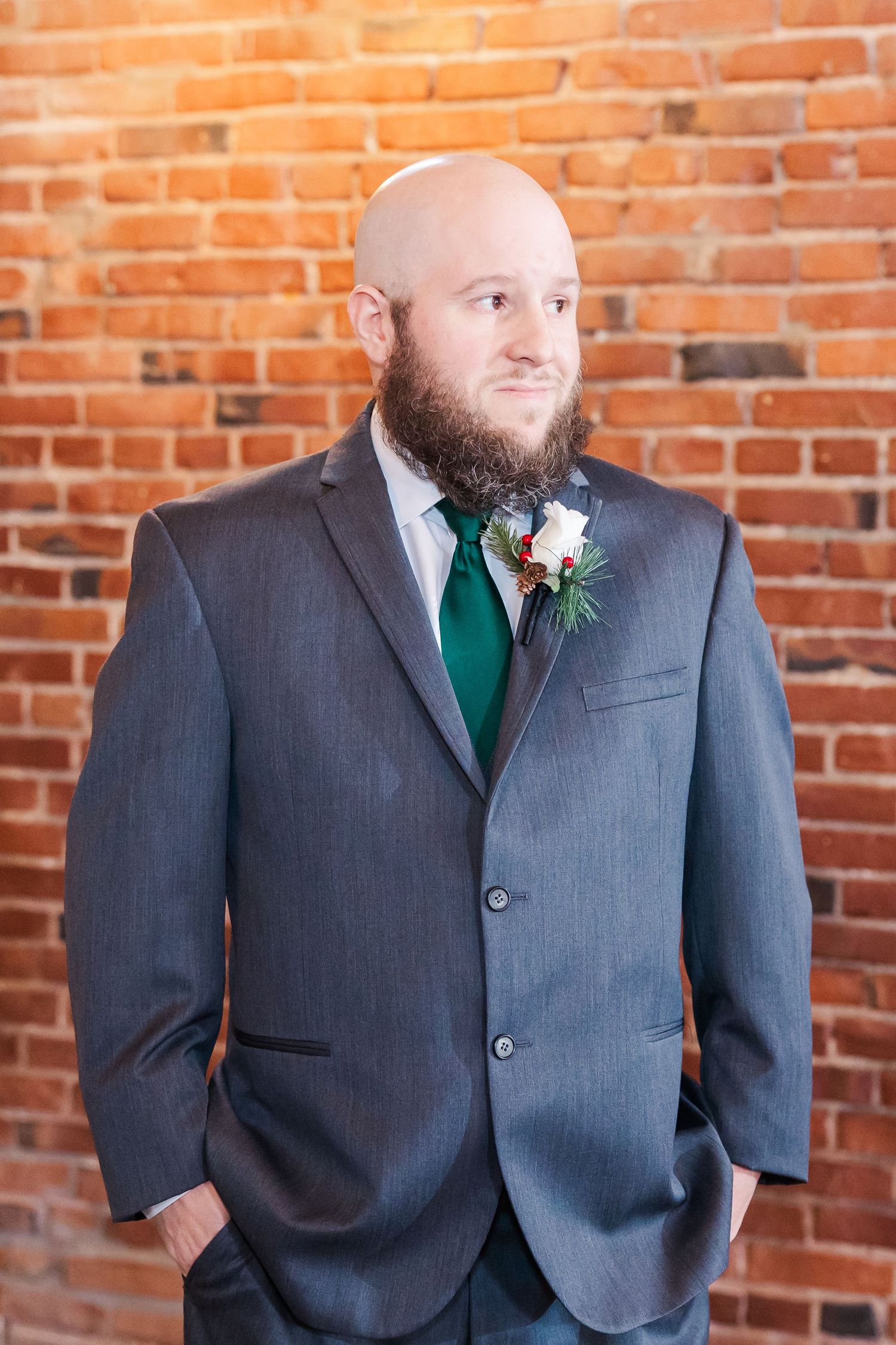 Groom in gray suit with emerald tie and boutonniere against brick wall.