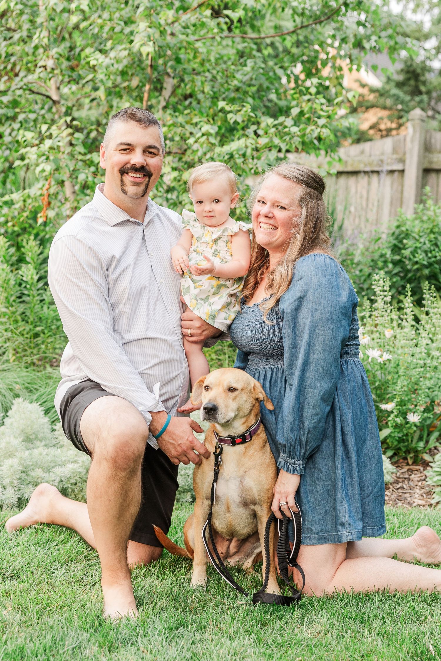 A loving family portrait outdoors with their golden retriever in a lush backyard garden.