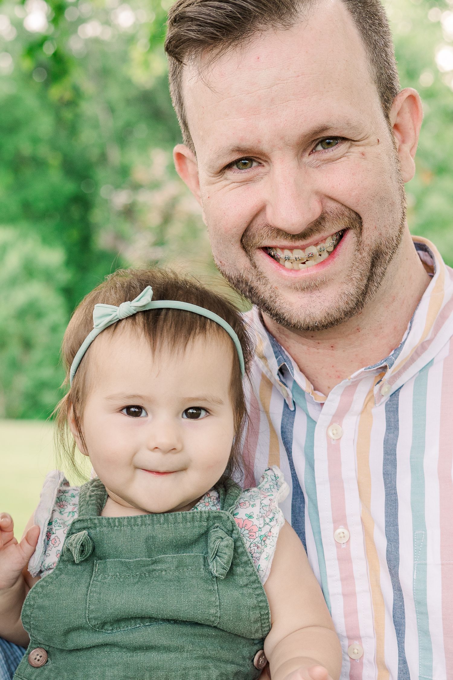 Portrait of a parent and young child in green overalls against a leafy background.