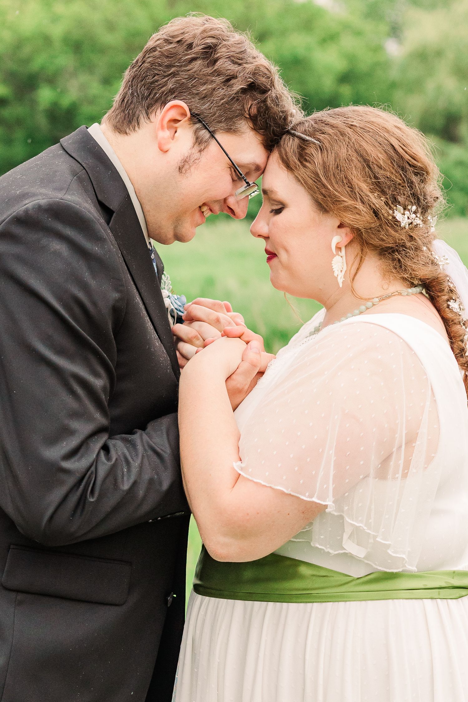 Intimate wedding portrait showing couple holding hands and touching foreheads in outdoor setting.
