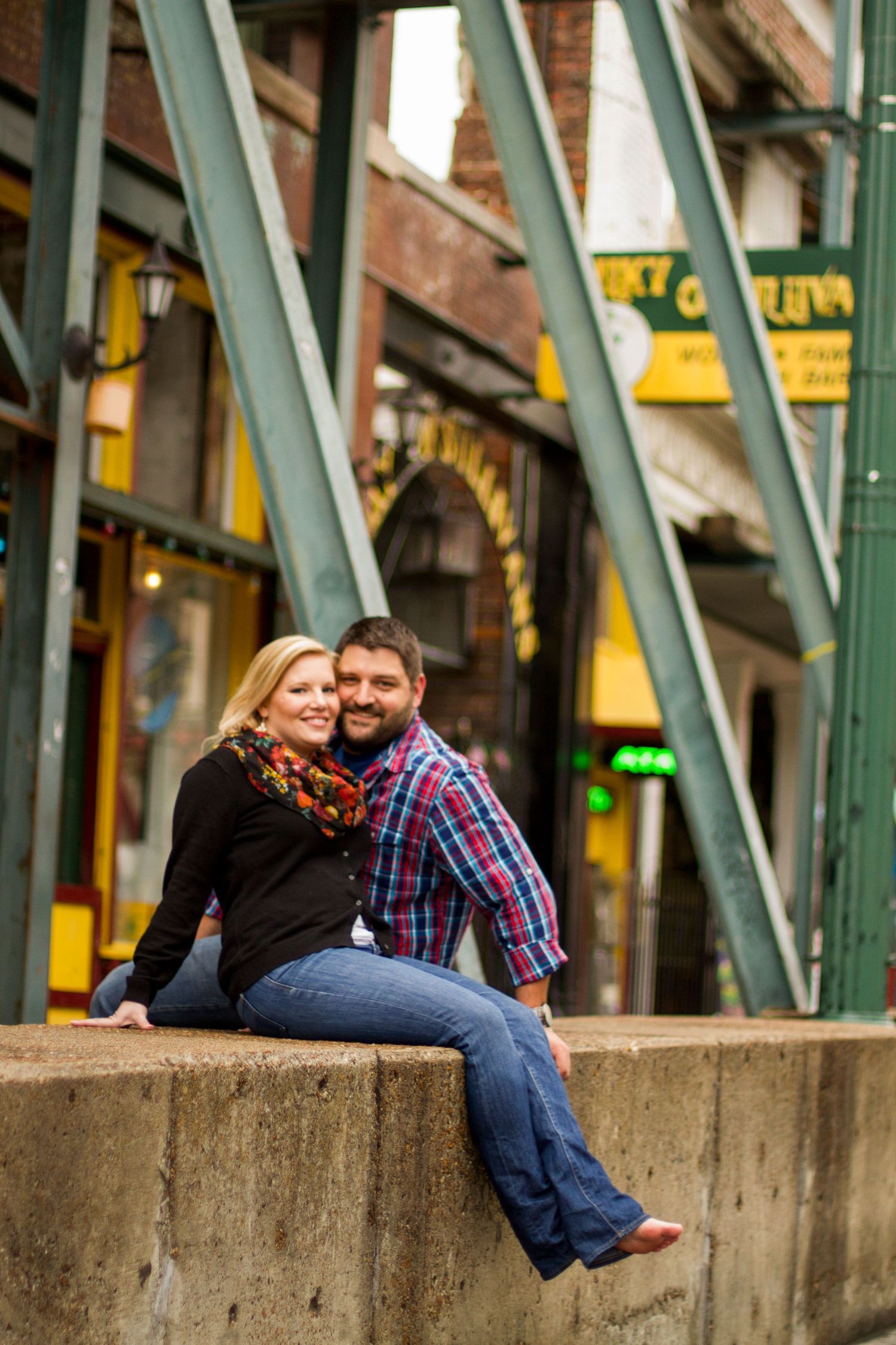 Engagement photo of couple sitting on concrete ledge with industrial backdrop.