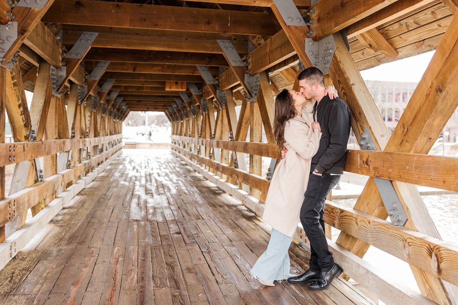 A couple shares a romantic moment inside a rustic covered bridge with wooden beams.