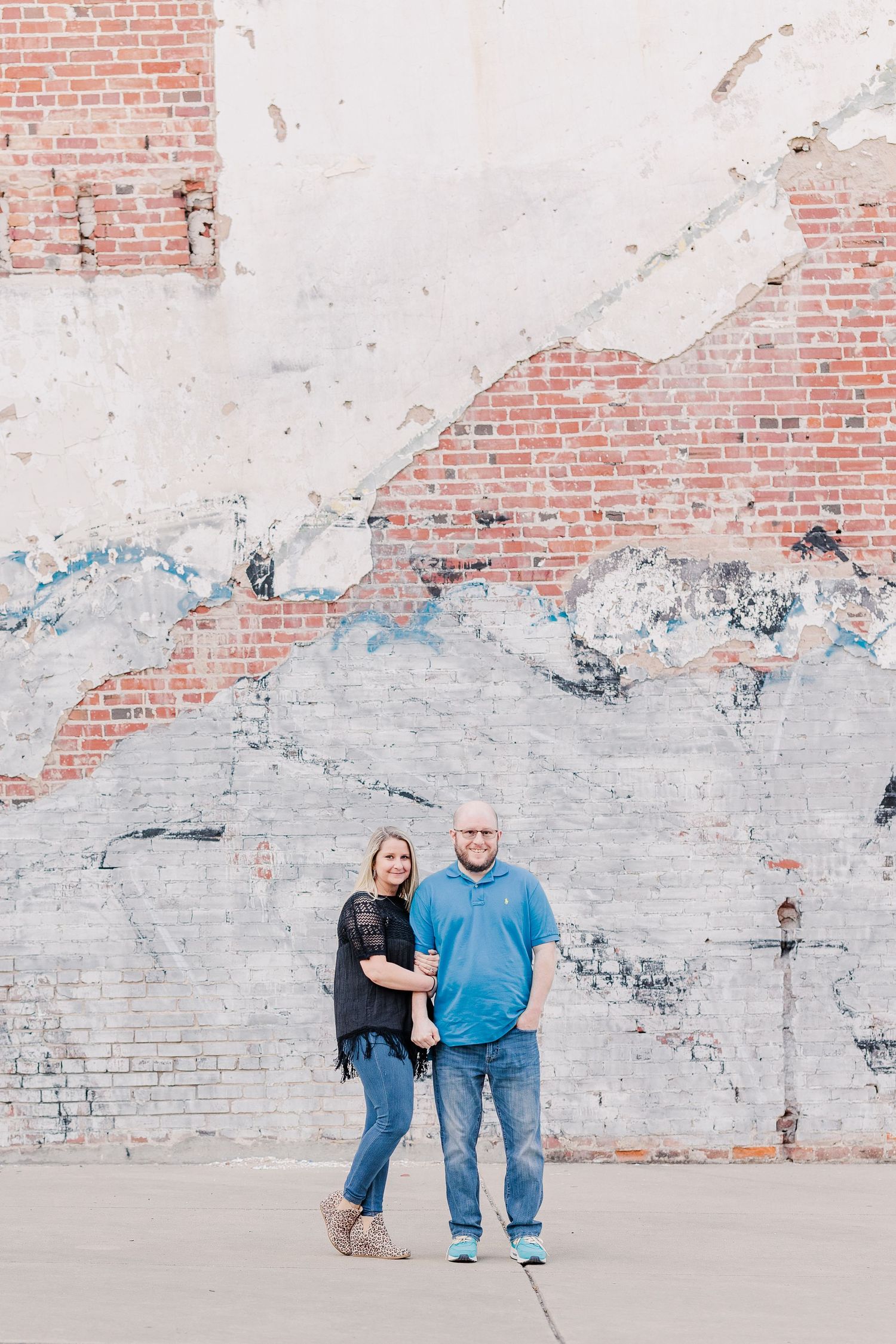 A couple stands together against a white and brick textured wall in casual attire.