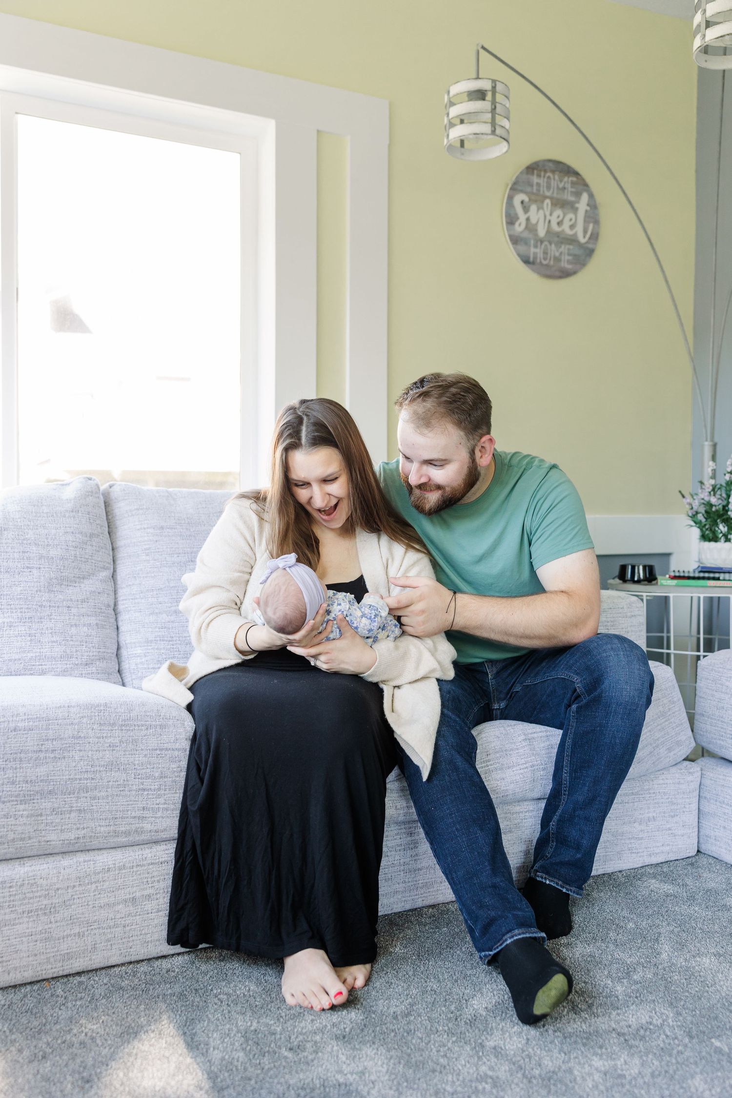 Happy couple sitting on gray sofa together admiring their newborn baby in living room.