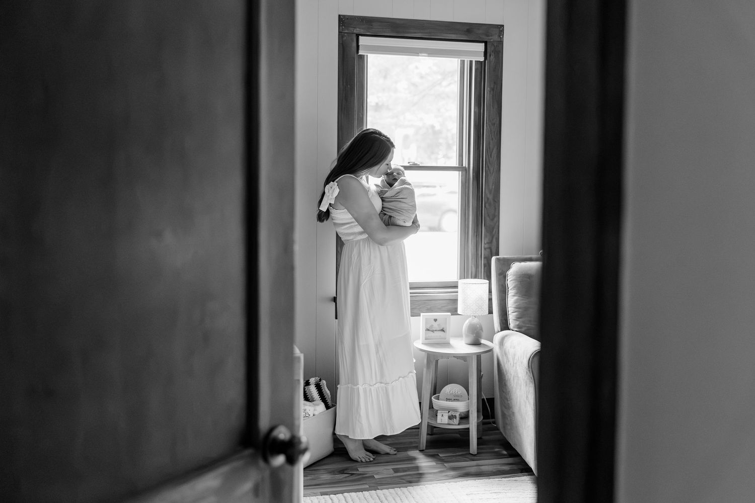 Black and white photo of a mother in a white dress cradling her newborn baby by a sunlit window.