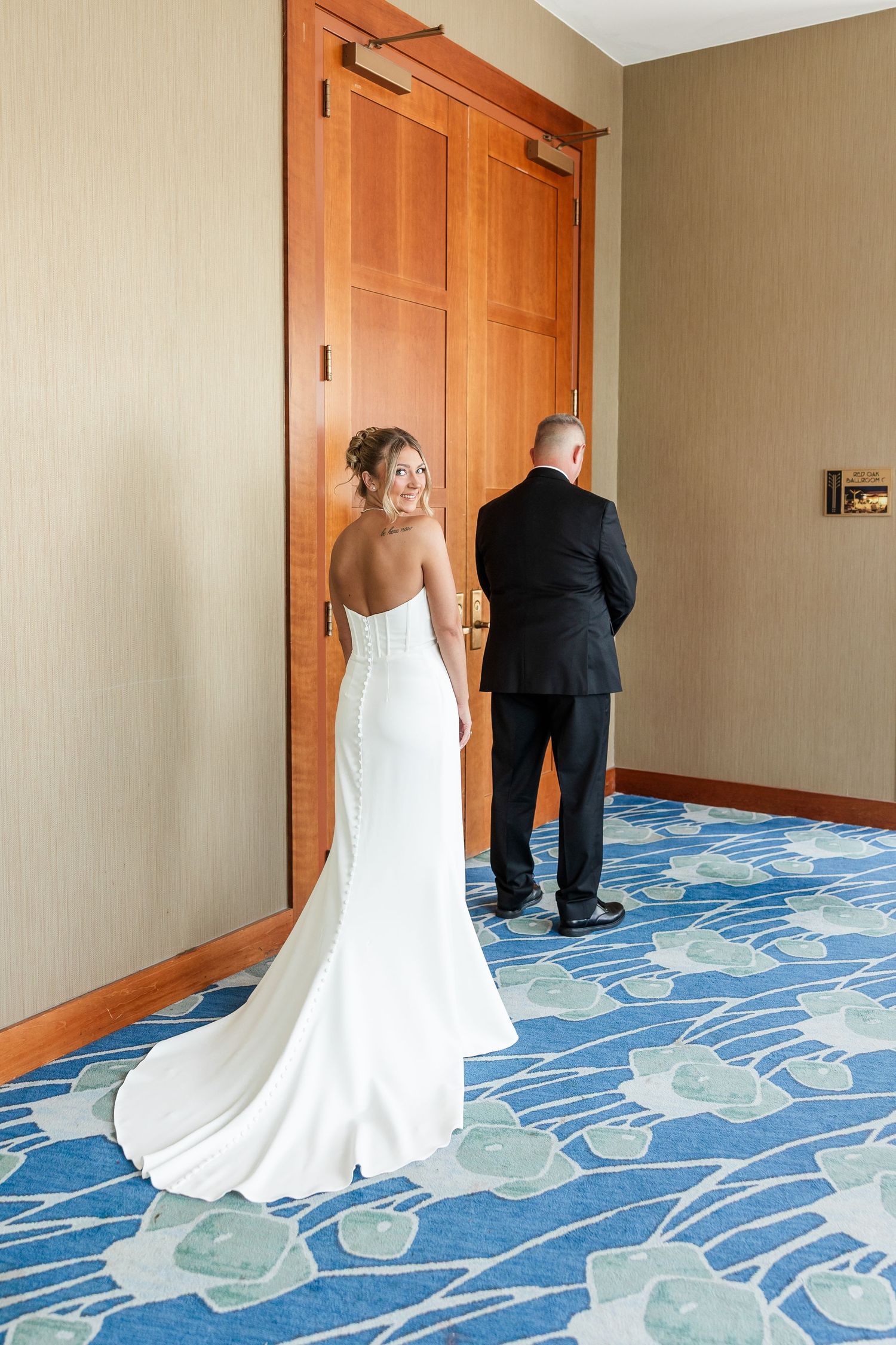 Bride in elegant white strapless gown with train approaches groom for first look in hotel hallway.