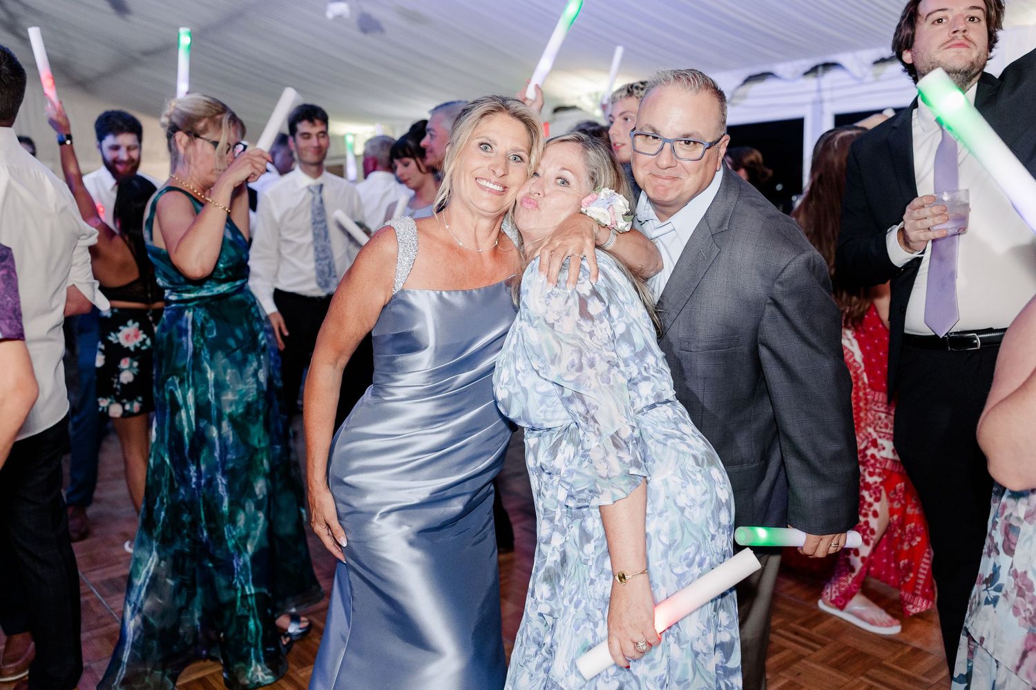 Guests celebrating at wedding reception dance floor holding glow sticks under white tent at night.