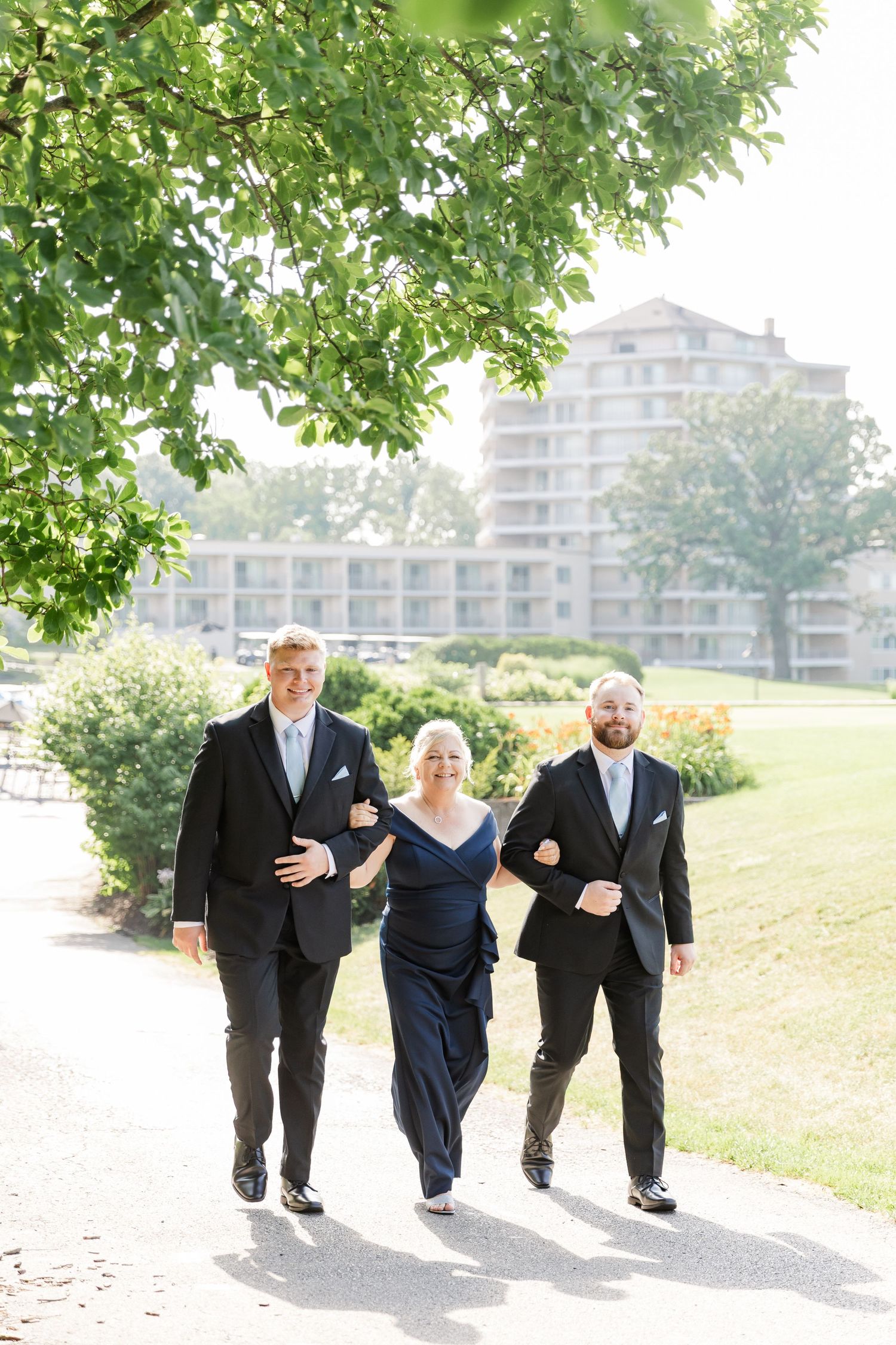 Groom and groomsman escort woman in navy dress along garden path at wedding venue.