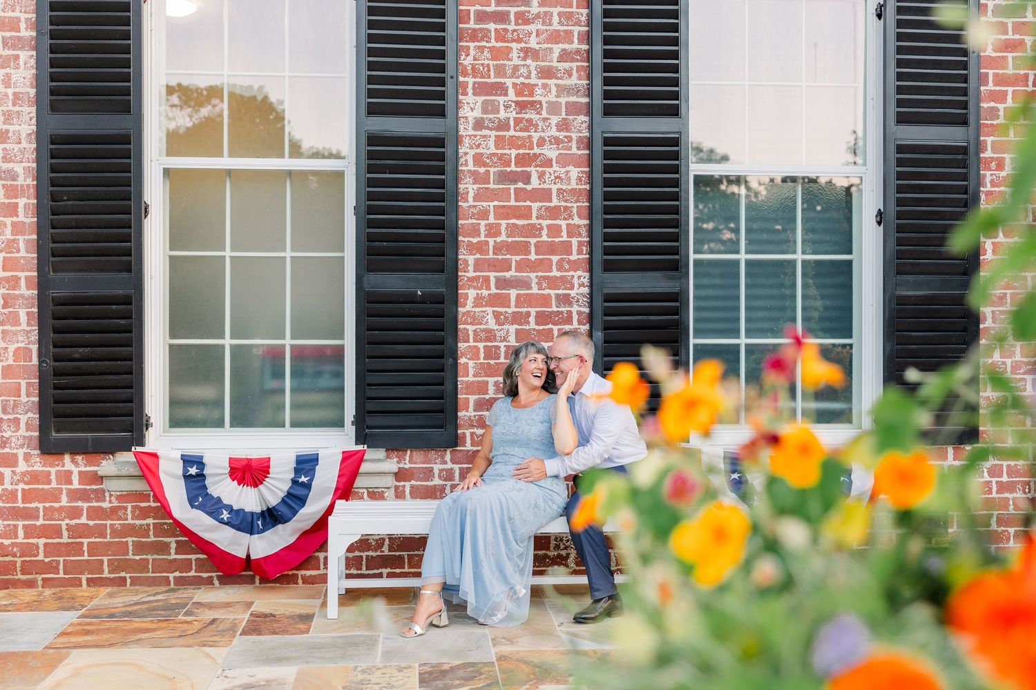 A smiling couple sits together on a bench outside a charming brick building adorned with colorful flowers.
