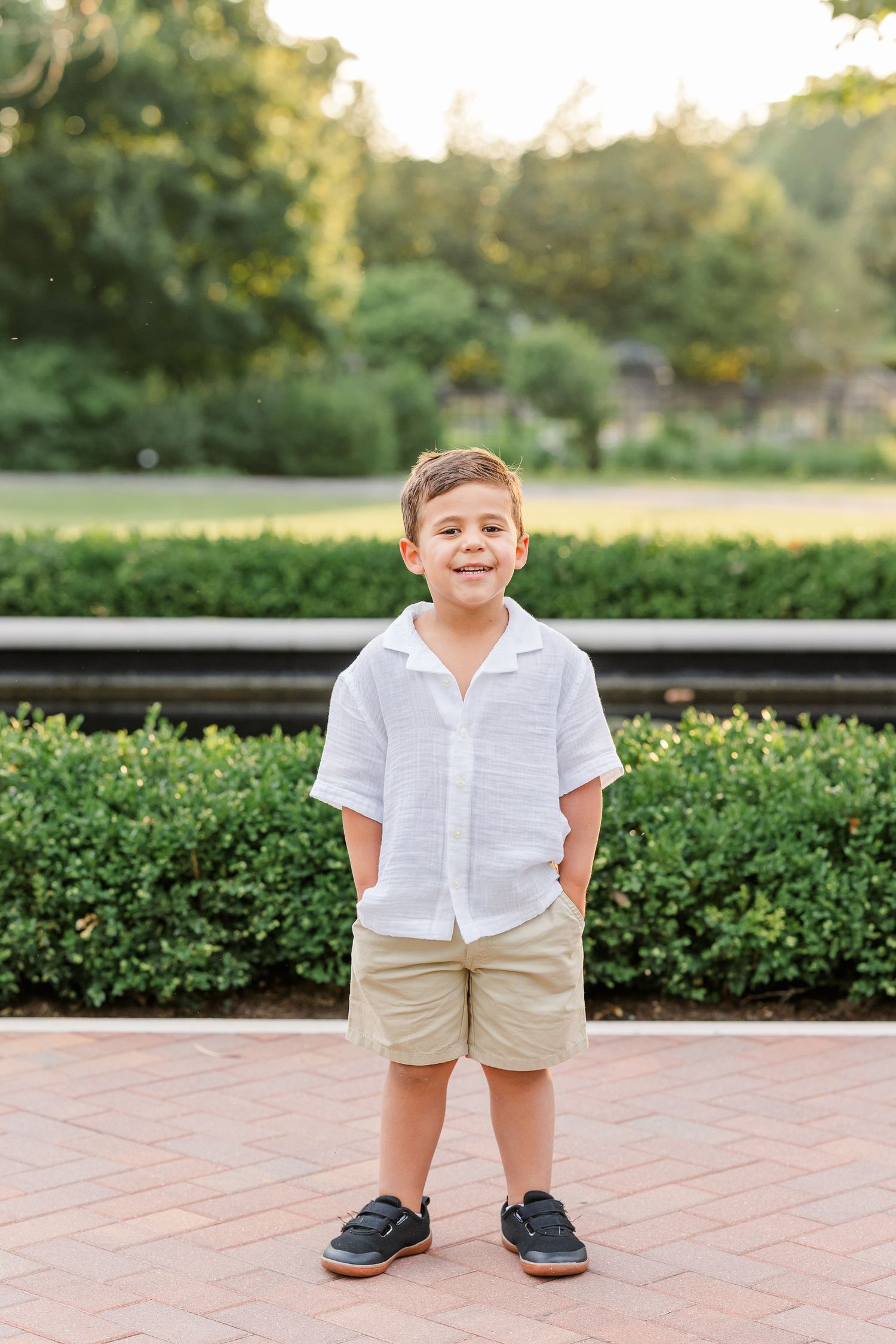 Young boy in white linen shirt and khaki shorts smiling outdoors in a garden setting.