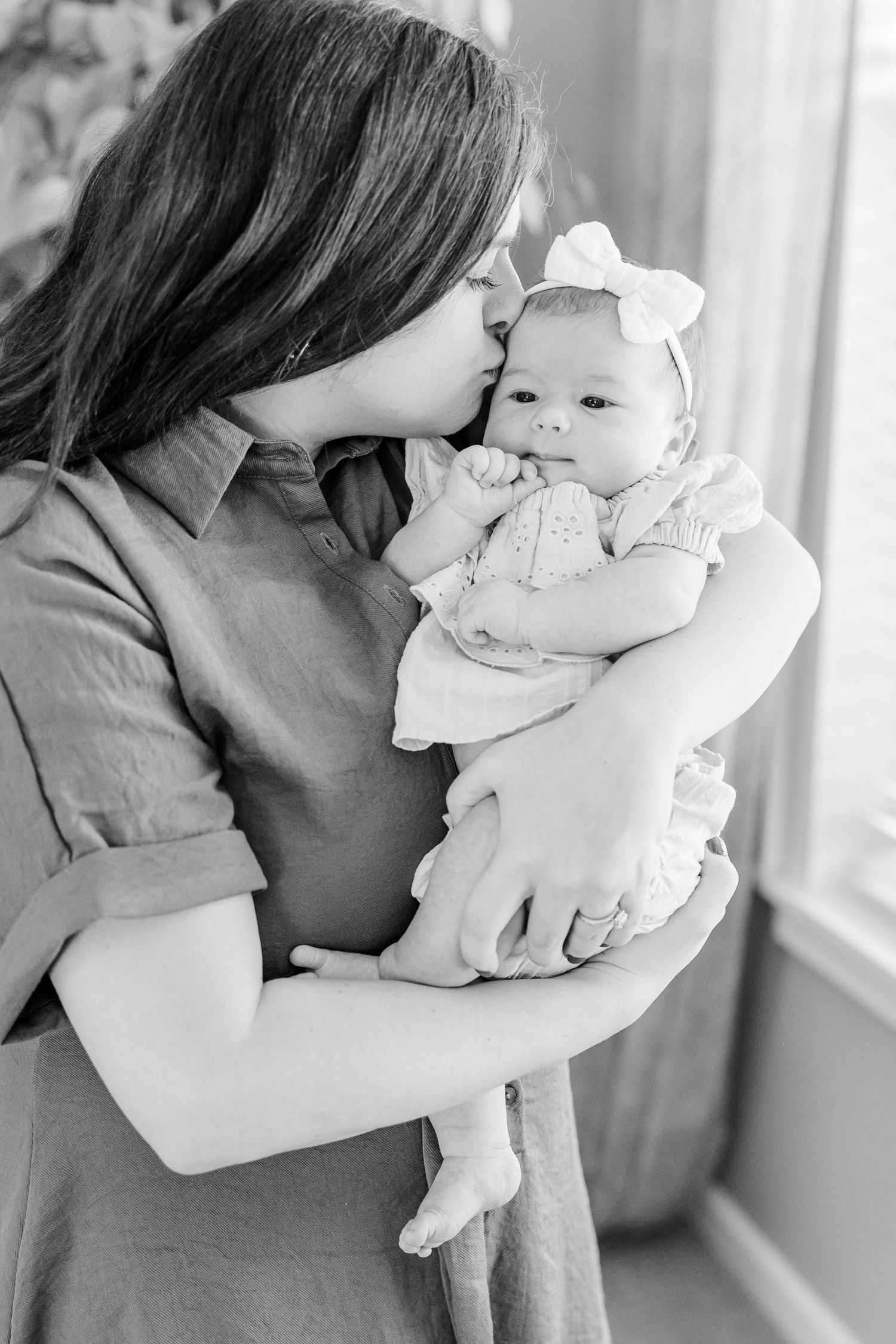 Black and white newborn photo of mom snuggling baby girl by the window during an in-home newborn session