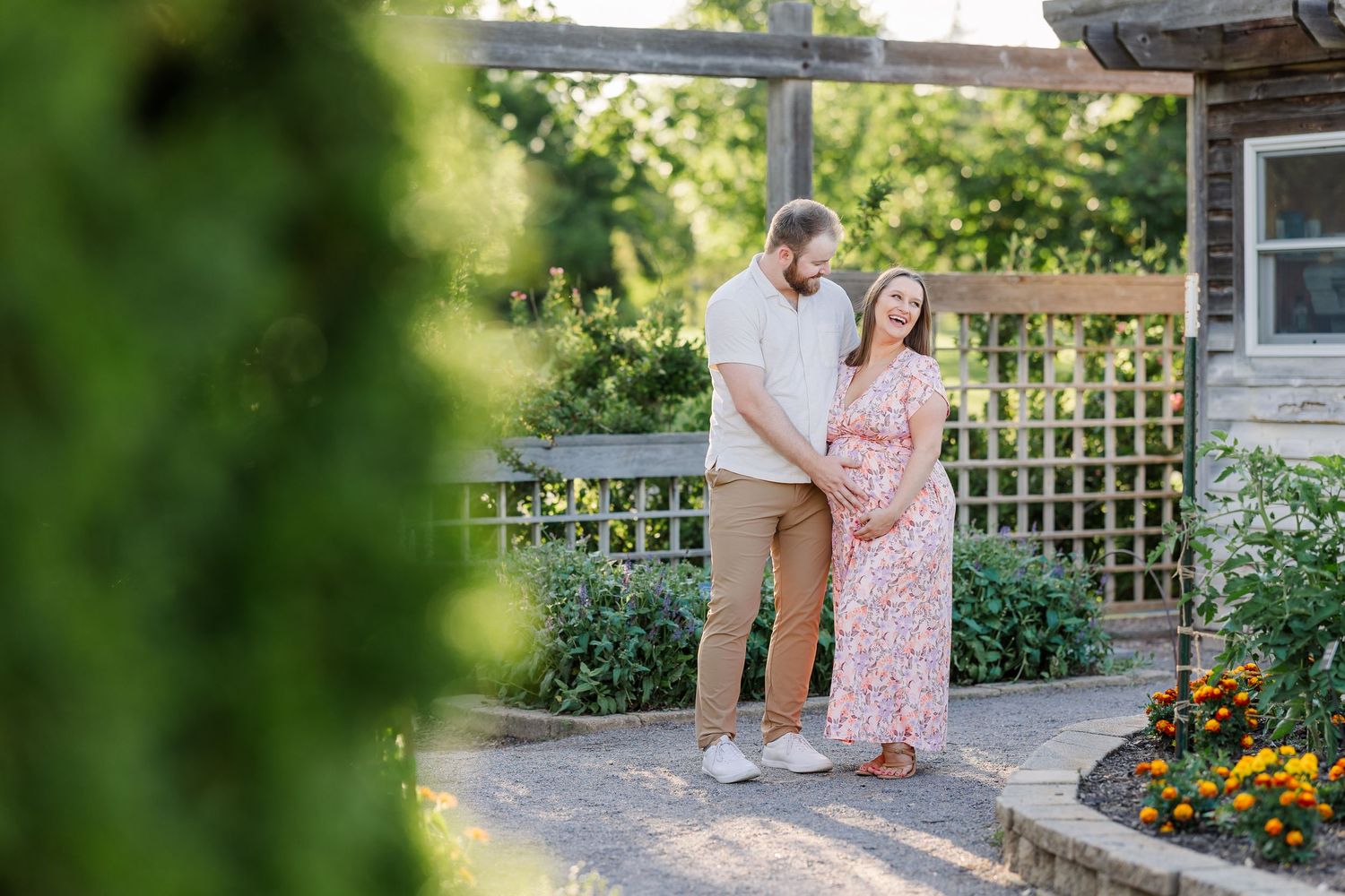 Couple posing in a lush garden, woman in floral dress cradling baby bump, man embracing her tenderly near wooden trellis.