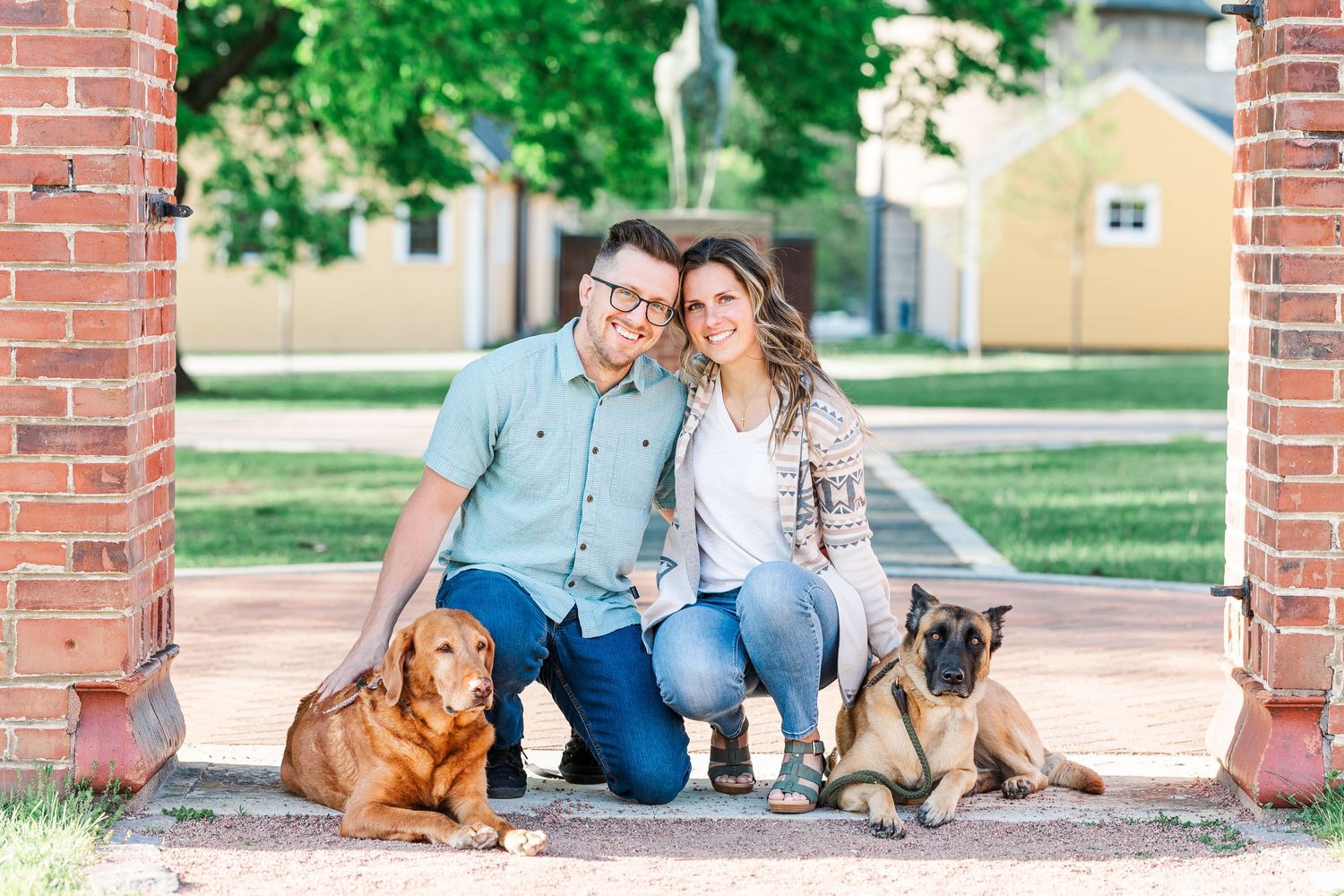 Engaged couple sitting with their dogs beneath the brick arch at St. James Farm Preserve
