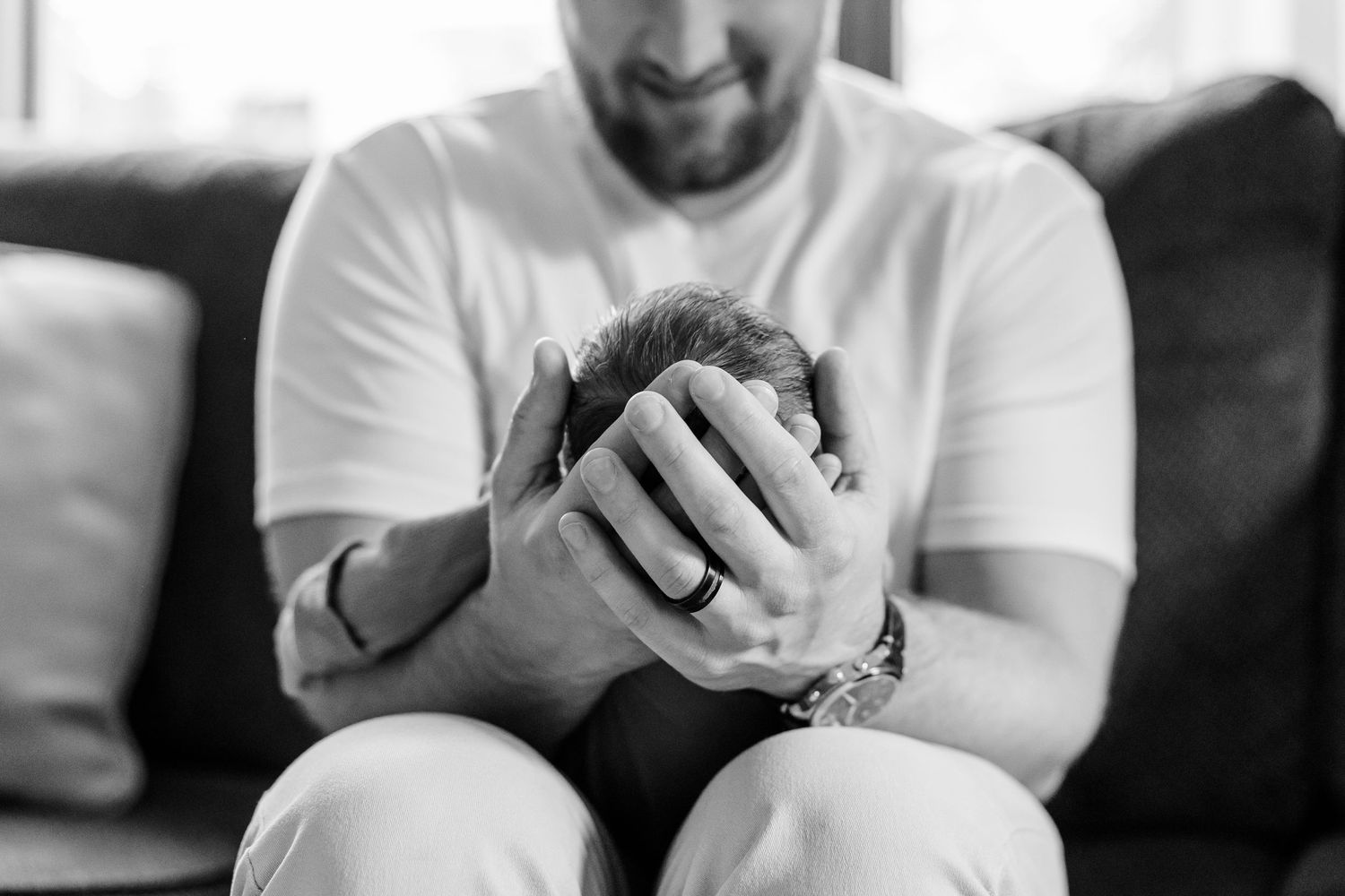 Black and white photo of a smiling bearded man cradling a newborn baby in his hands while seated on a couch.
