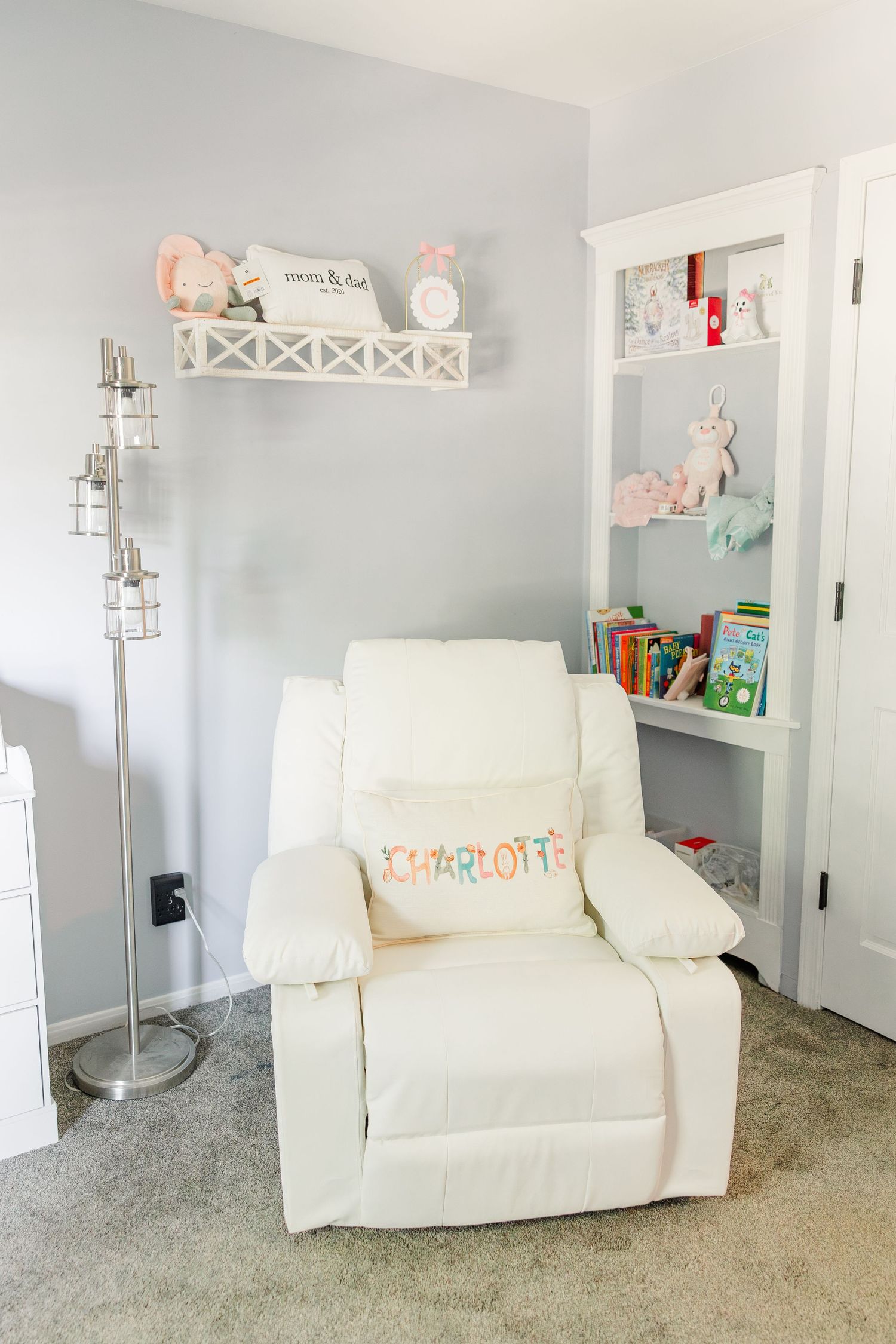 White nursery recliner chair with Charlotte name pillow beside bookshelf and wall shelf with toys.