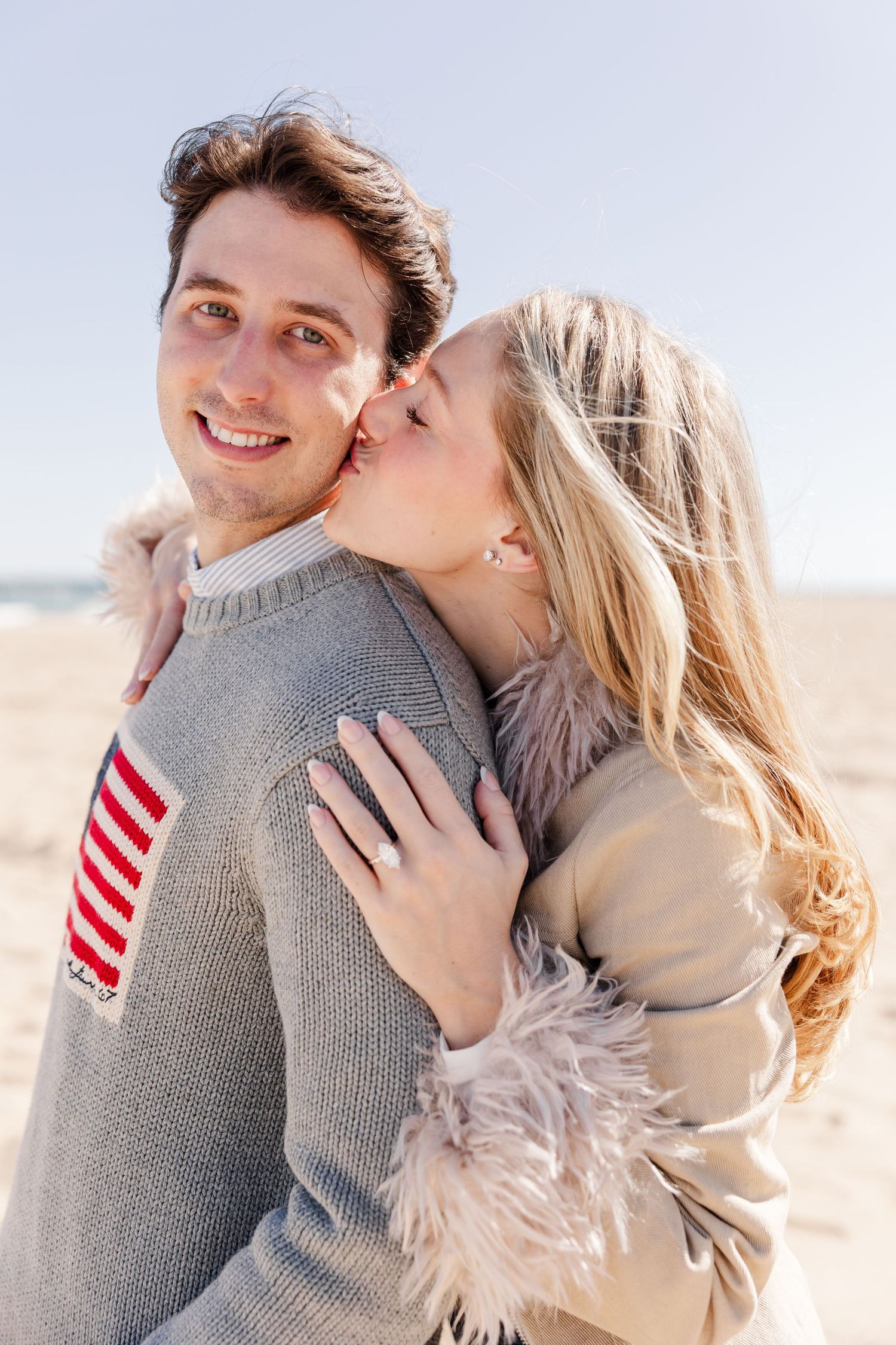 A happy couple embraces on a beach, with the woman kissing the smiling man on the cheek while showing an engagement ring.