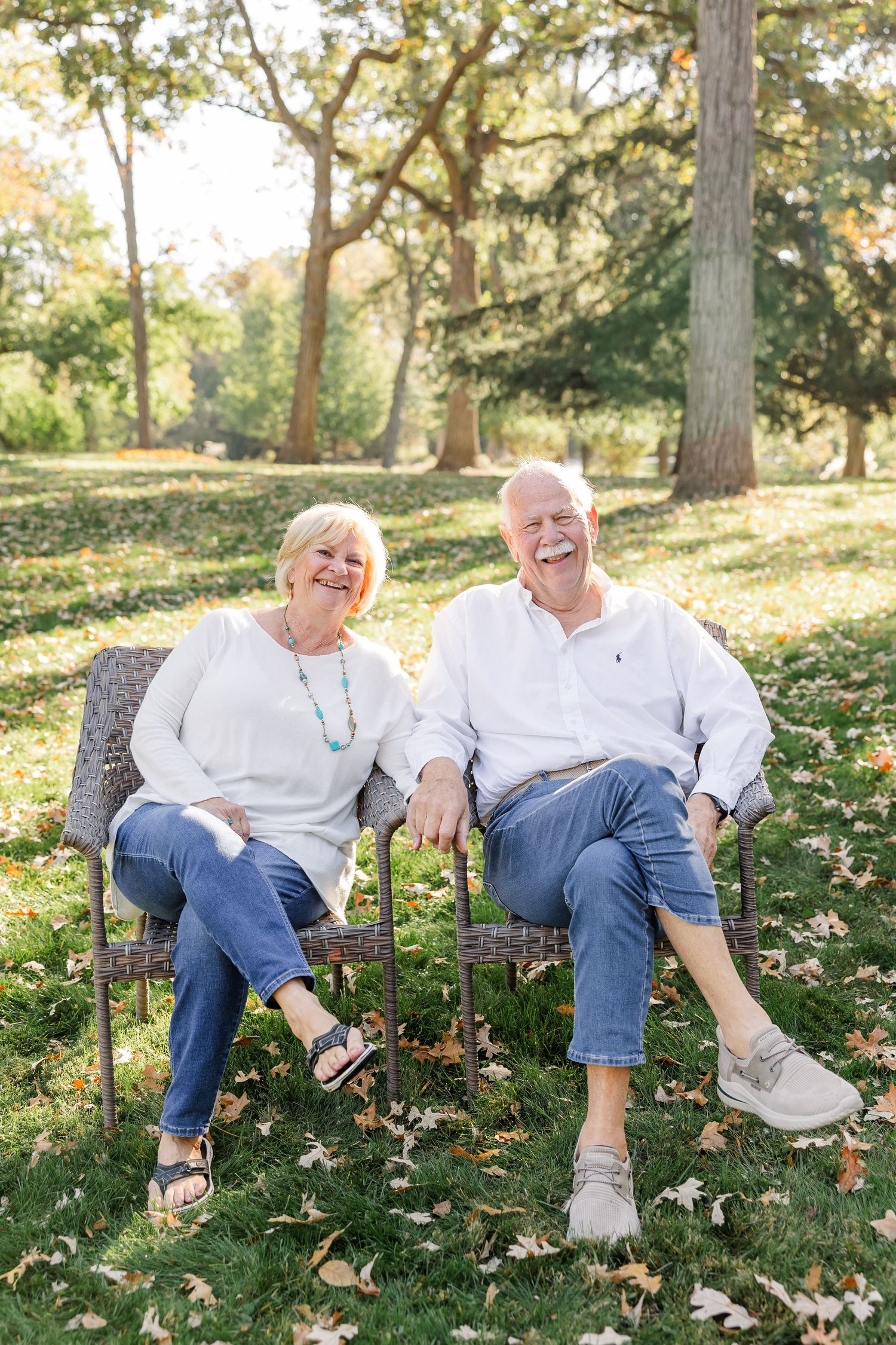 Elderly couple laughing joyfully while sitting on rustic chairs in a park during autumn.