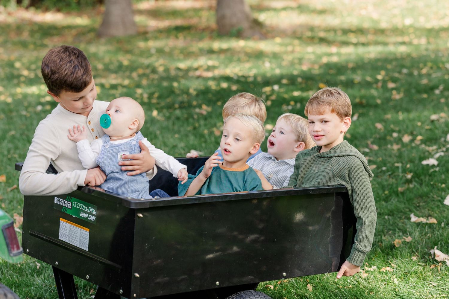 Five young children sit together in a black wagon on green grass in a sunny park setting.